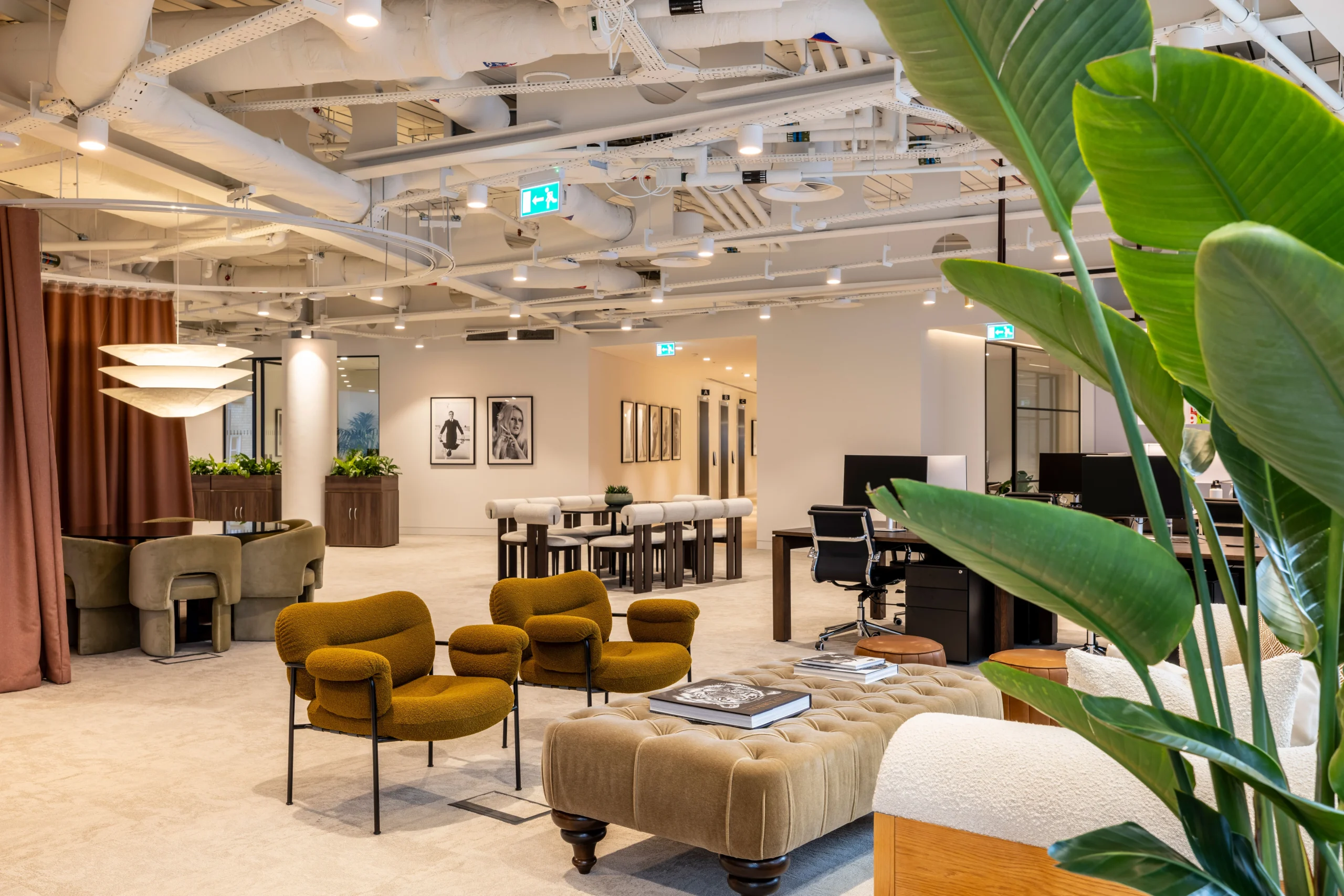 Authentic Brands Group workspace, featuring an open-plan design with stylish furniture, including mustard armchairs, a tufted ottoman, and a mix of collaborative and desk seating. The space is accented with greenery, framed artwork, and exposed ceiling details.