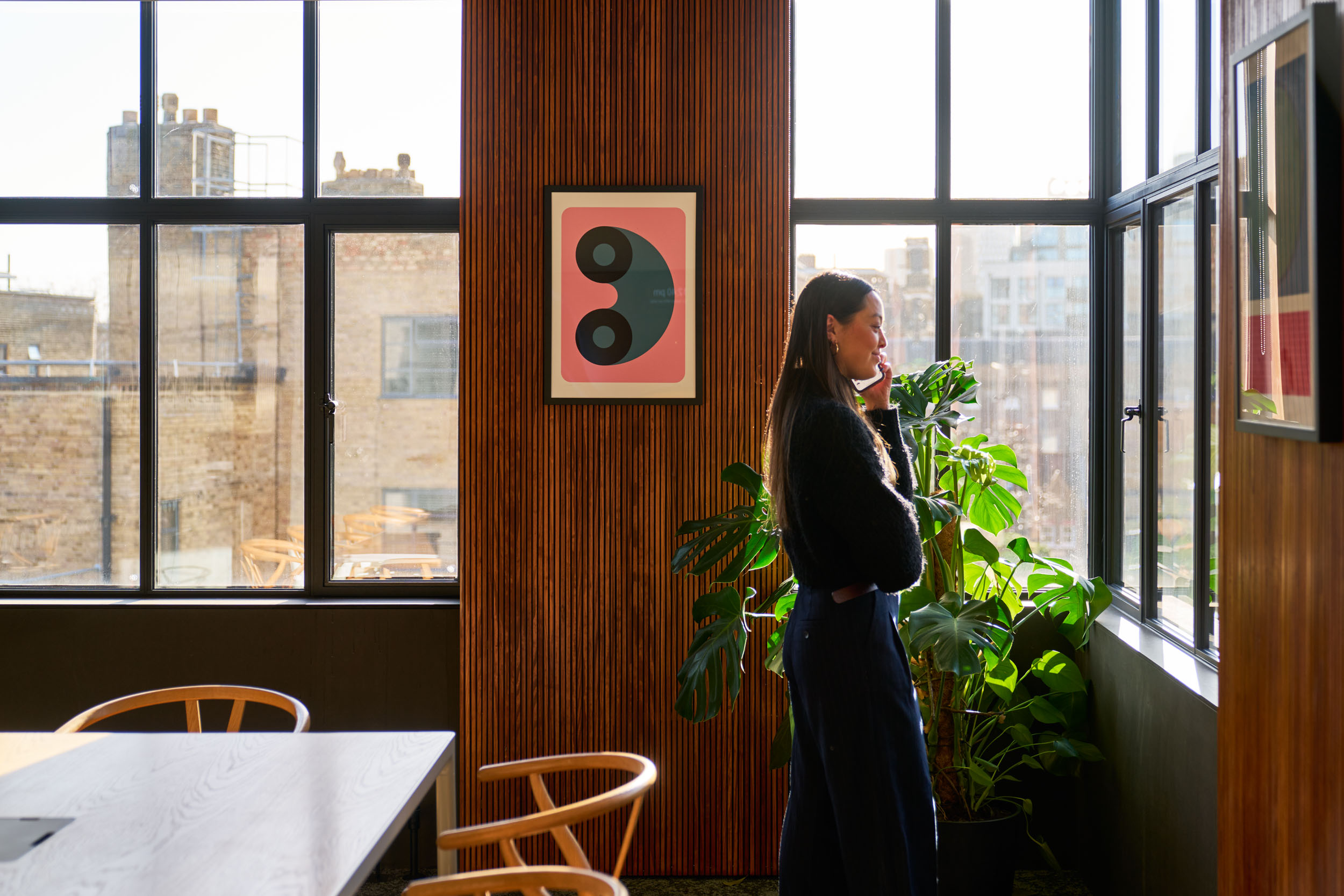 Person standing by a window on the phone in Joseph Joseph's workspace, featuring wooden slat panelling, lush green plants, and framed artwork, capturing a modern and functional design aesthetic.