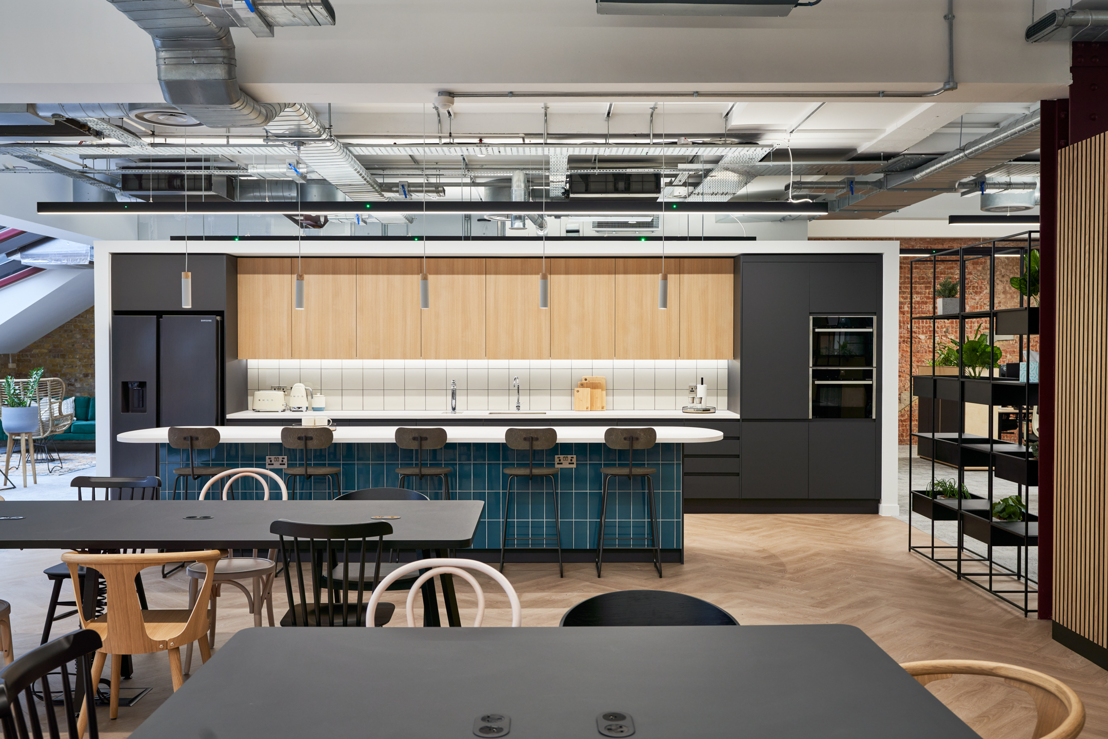A modern office kitchen with sleek wood paneling, white countertops, and teal bar stools providing a pop of color. The space is lit with suspended linear lights and features advanced kitchen appliances, embodying a blend of functionality and style.