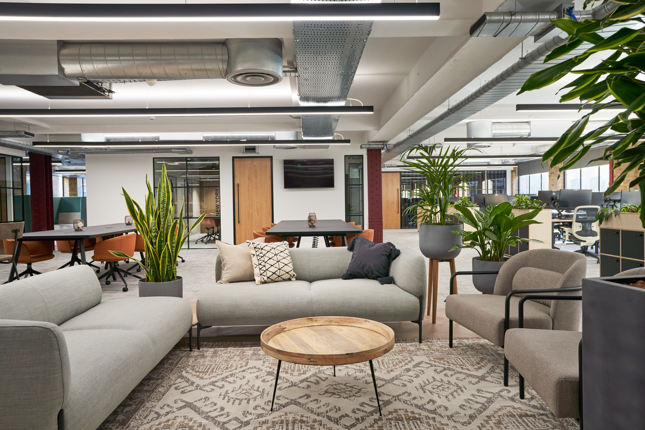 A hybrid lounge and work area furnished with soft grey sofas and chic armchairs, complemented by lush plant decor and a large wooden coffee table. The space is designed to facilitate relaxation and casual work, with a homely yet professional atmosphere.