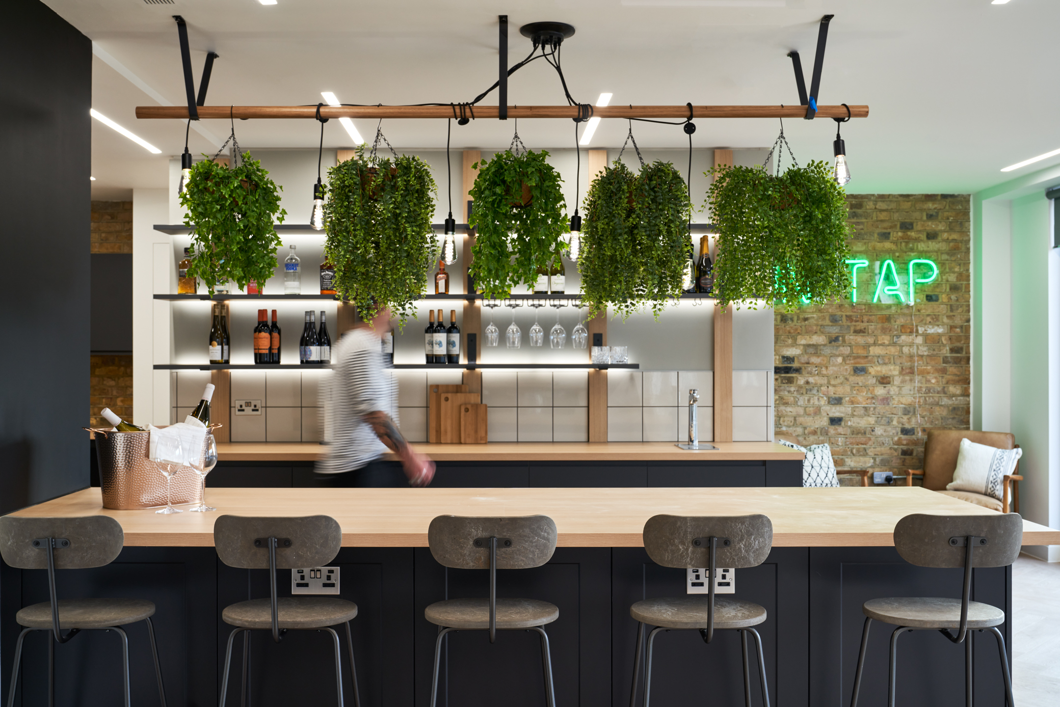 A lively office bar area featuring hanging greenery that adds vibrancy and a touch of nature. The bar is equipped with modern facilities and stylish seating, making it ideal for informal meetings or breaks.