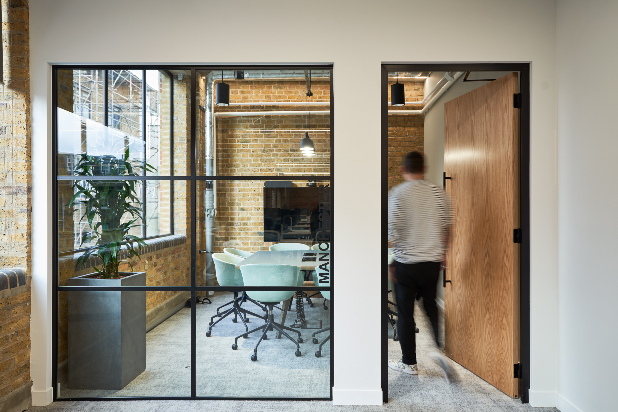 A glass-walled meeting room showcasing a clean, modern design with exposed brick and light wood accents. This setting provides transparency while maintaining a sense of privacy and is equipped with contemporary office furniture.
