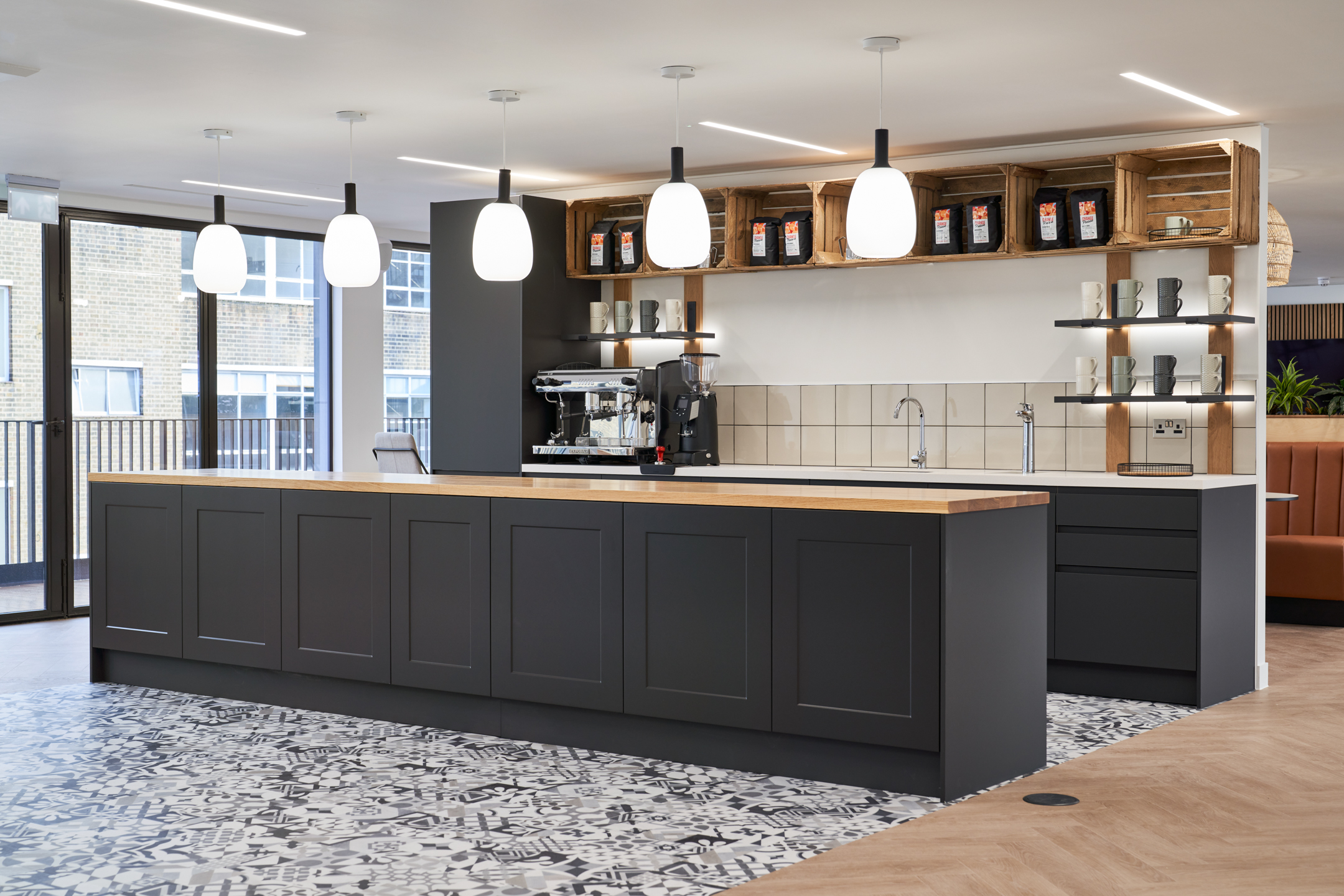 Ayden's chic barista station in a corporate setting, featuring dark grey cabinetry and a professional espresso machine. The area is decorated with white hanging lamps and rustic wooden crates, creating a warm and inviting atmosphere for coffee breaks.