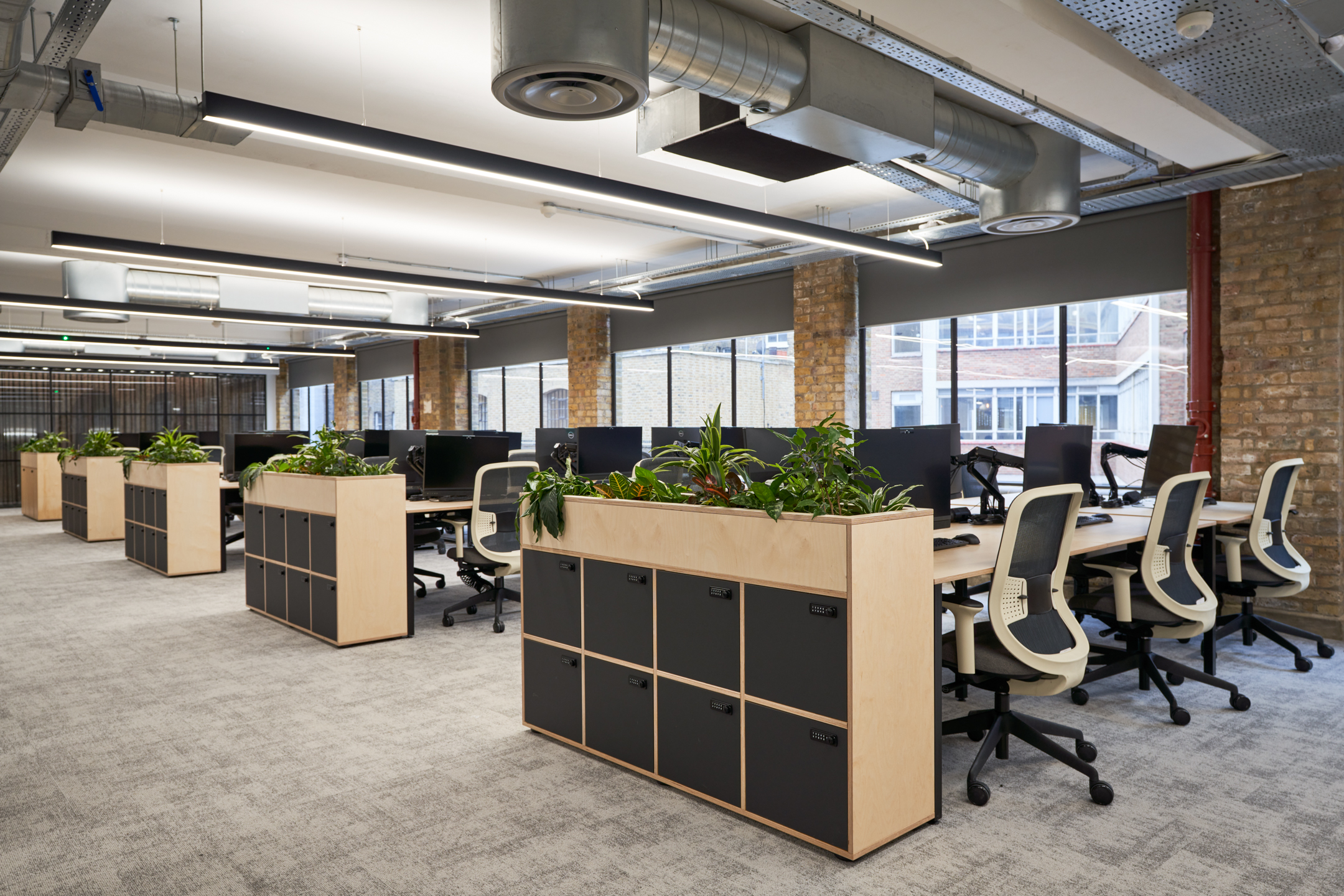 A spacious open-plan office featuring light grey carpeting and modular storage planters, serving both aesthetic and practical purposes. The office is equipped with ergonomic chairs and ample natural light, providing a comfortable working environment.