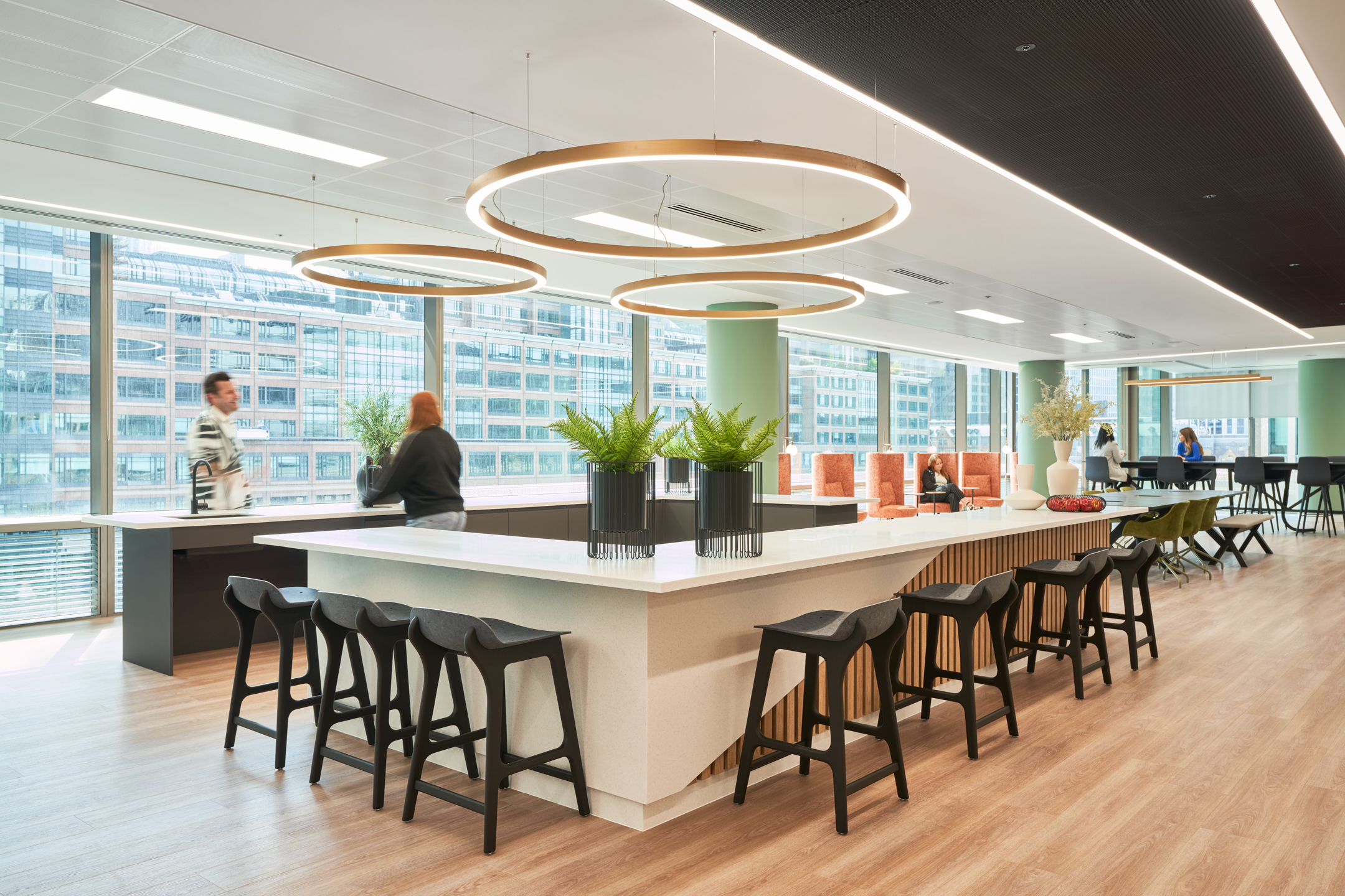 A modern office kitchen area at British Land's 10 Exchange Square, designed by Modus Workspace, featuring sleek countertops, black stools, and circular light fixtures. Two people are seen interacting in the bright, open space.