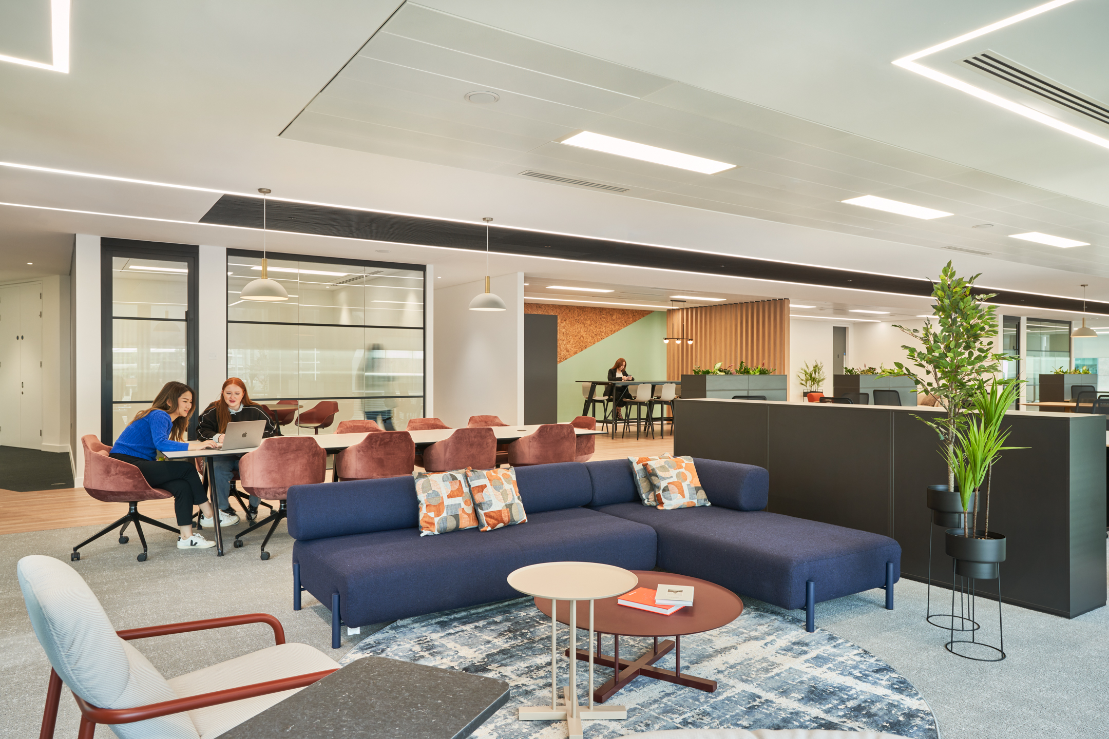 A vibrant office lounge area at British Land’s 10 Exchange Square, designed by Modus Workspace, with blue sectional sofas, red chairs, and collaborative workspaces. Two people are engaged in discussion while another works in the background.