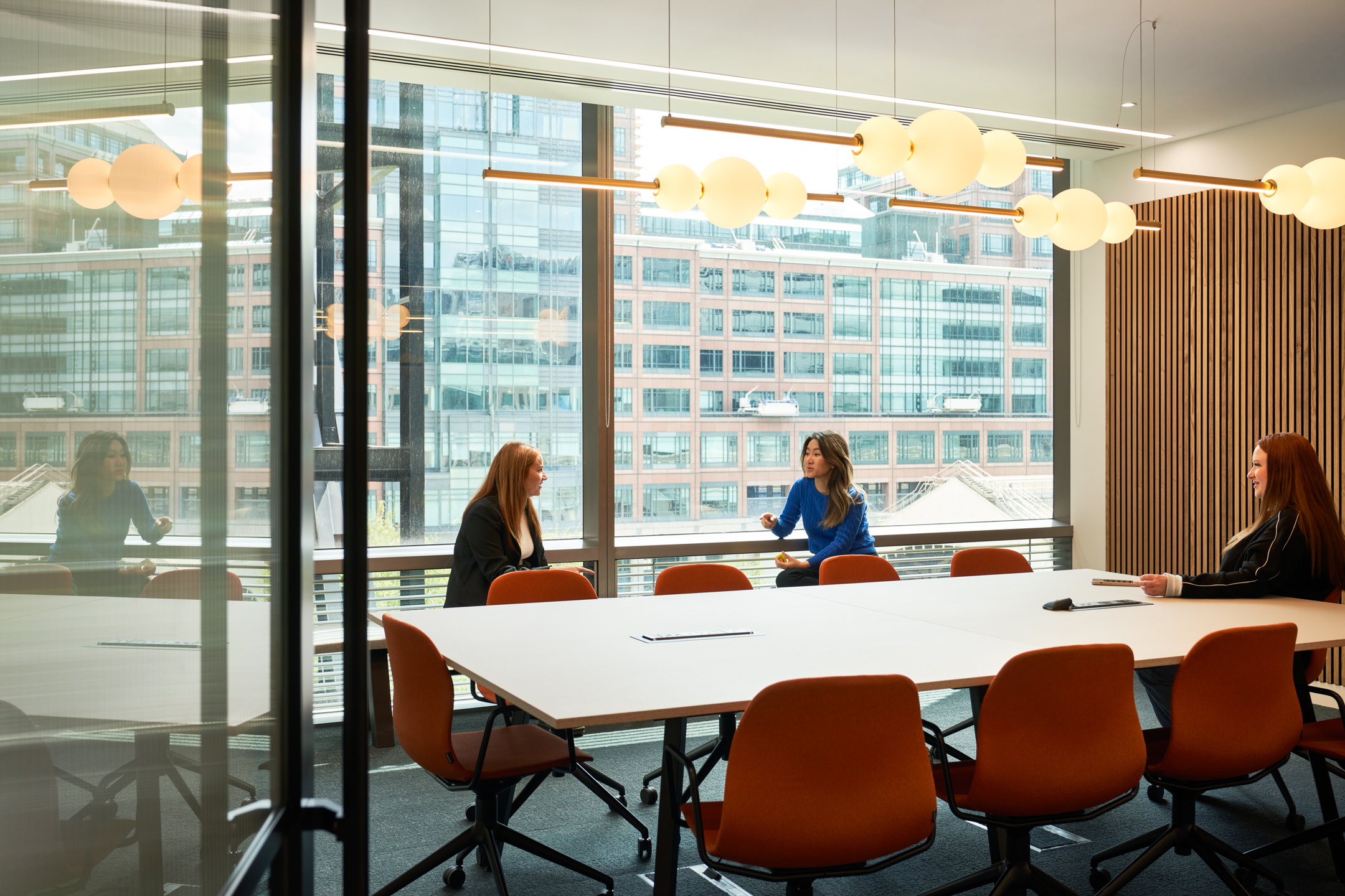 A glass-enclosed meeting room at British Land's 10 Exchange Square, designed by Modus Workspace, with orange chairs and large windows offering city views. Three people are seated around the table in discussion