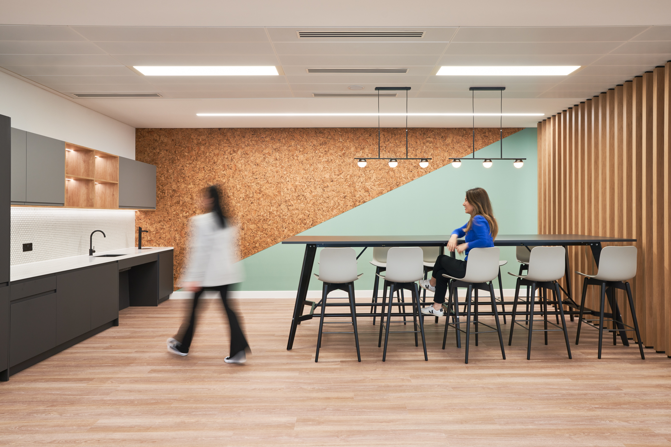 A modern teapoint and break-out space at British Land’s 10 Exchange Square, designed by Modus Workspace, with a cork accent wall and mint green details. One person is sitting at a high table while another walks by.