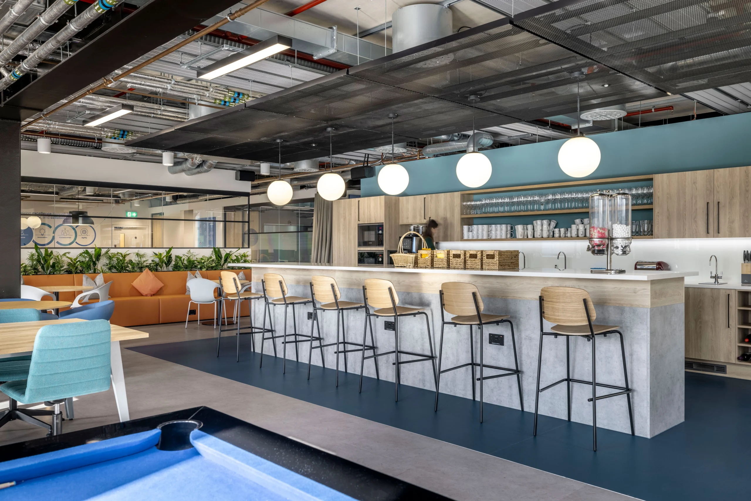 A modern teapoint designed by Modus Workspace for Carbon Clean, featuring a long counter with bar stools, overhead pendant lights, and an open ceiling exposing industrial pipes. The space includes a mix of seating options and greenery for a vibrant and collaborative environment.