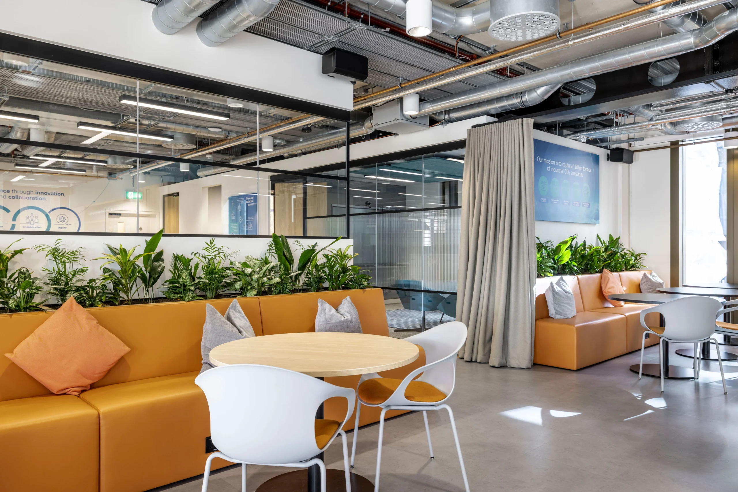 A comfortable seating area in Carbon Clean's office designed by Modus Workspace, showcasing a long orange bench with accent pillows, round tables with white chairs, and integrated greenery. The design creates a relaxing space for informal meetings and breaks.