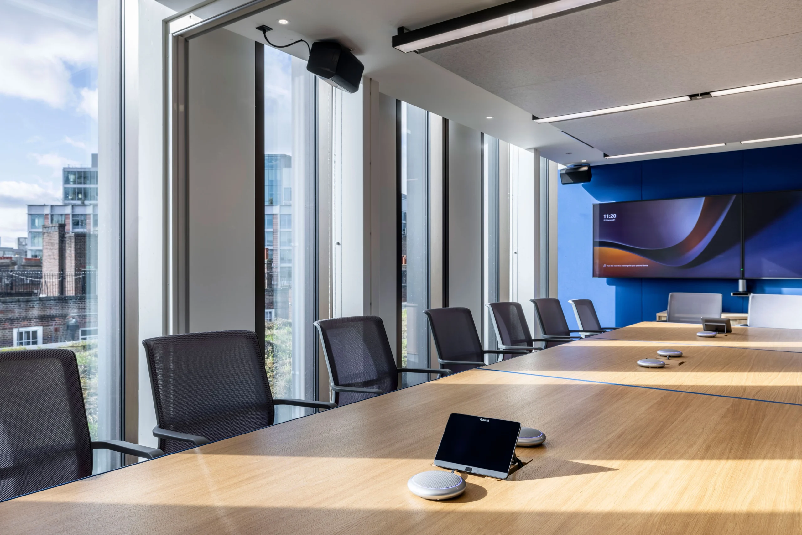A bright and modern meeting room at Carbon Clean's office, designed by Modus Workspace, featuring a large wooden conference table, ergonomic chairs, and floor-to-ceiling windows that offer plenty of natural light and views of the surrounding cityscape.