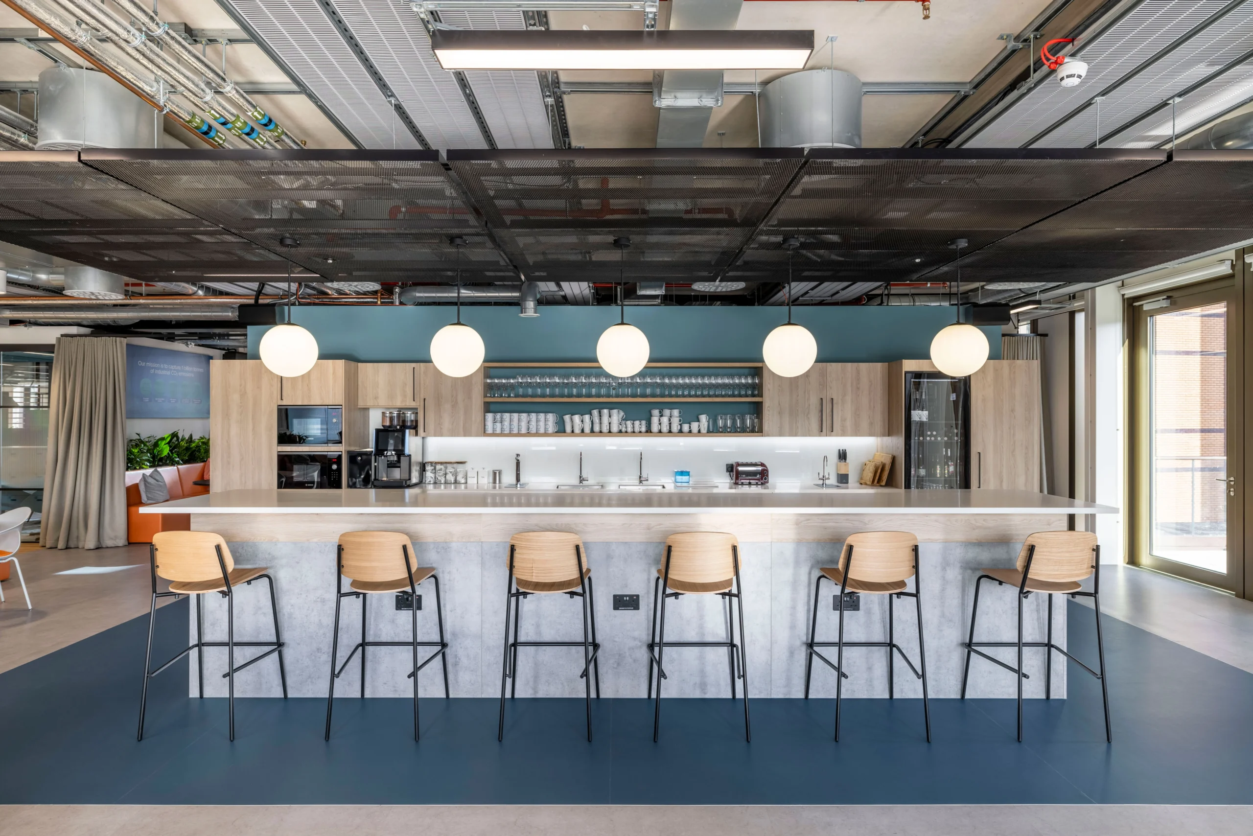 A view of the office teapoint in Carbon Clean's workspace, designed by Modus Workspace, with a focus on the clean and functional design of the kitchen counter, high stools, and pendant lights that create an inviting space for employees to gather.
