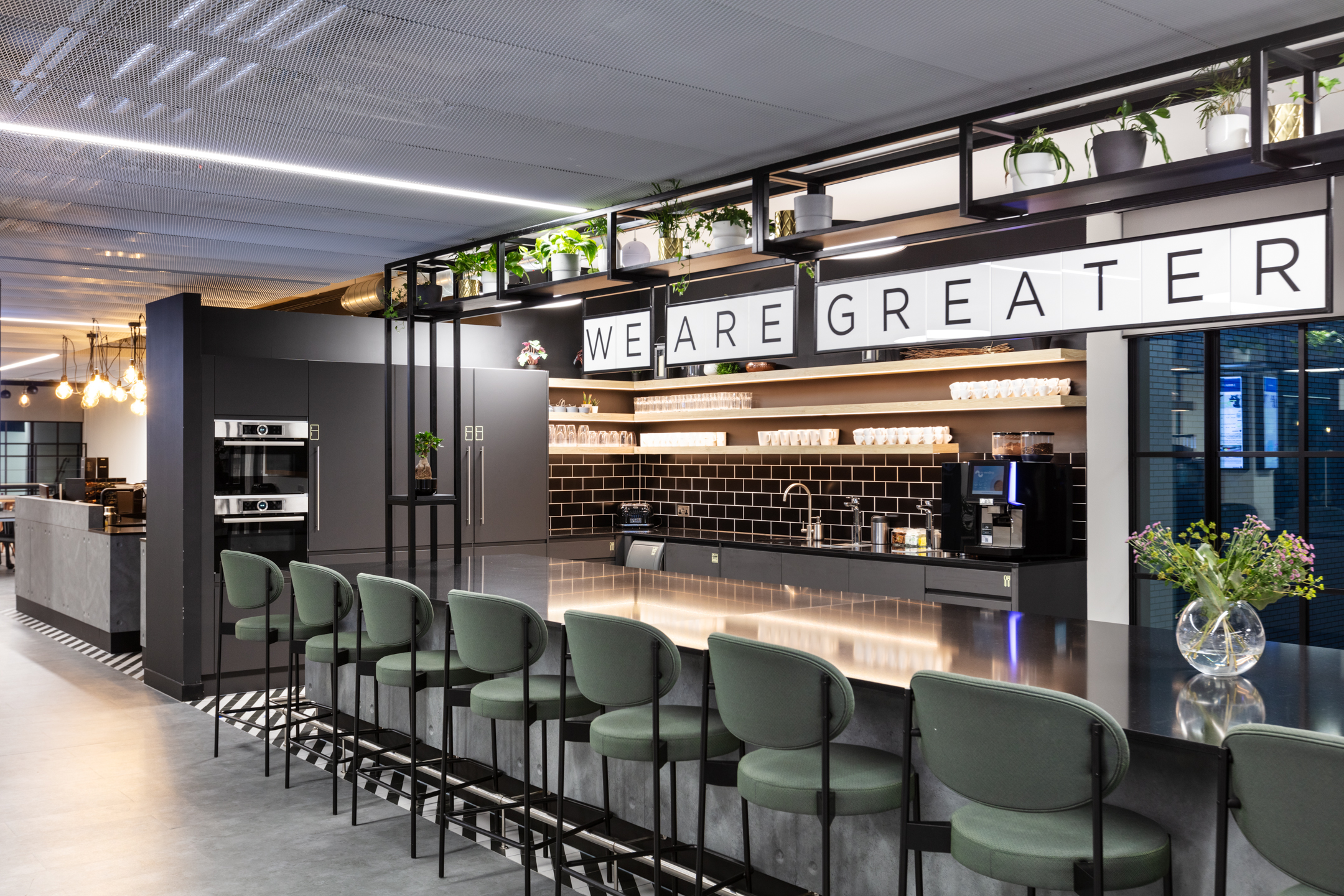 A stylish office kitchen and bar area featuring dark grey cabinetry, black subway tile backsplash, and a long counter with green high stools. Overhead, exposed industrial beams complement the modern light fixtures, and shelves adorned with plants and signage reading