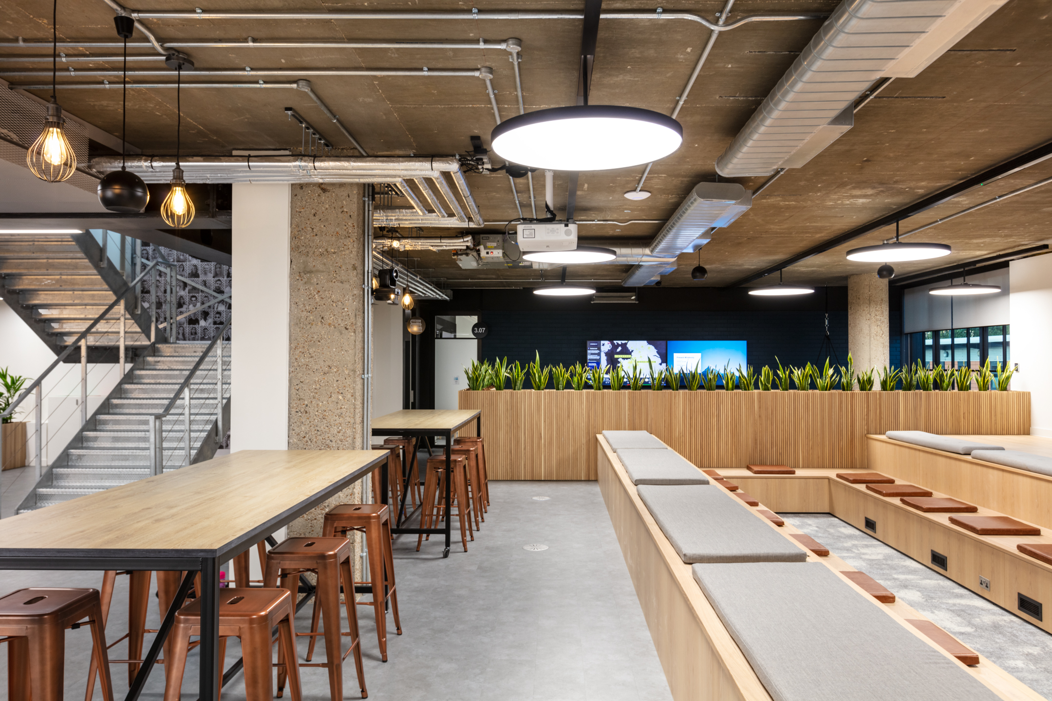 A spacious office area combining work and relaxation zones. The space includes high wooden tables with copper stools, and a comfortable seating area with long benches. Exposed ceilings and strategic lighting provide an urban, contemporary look.