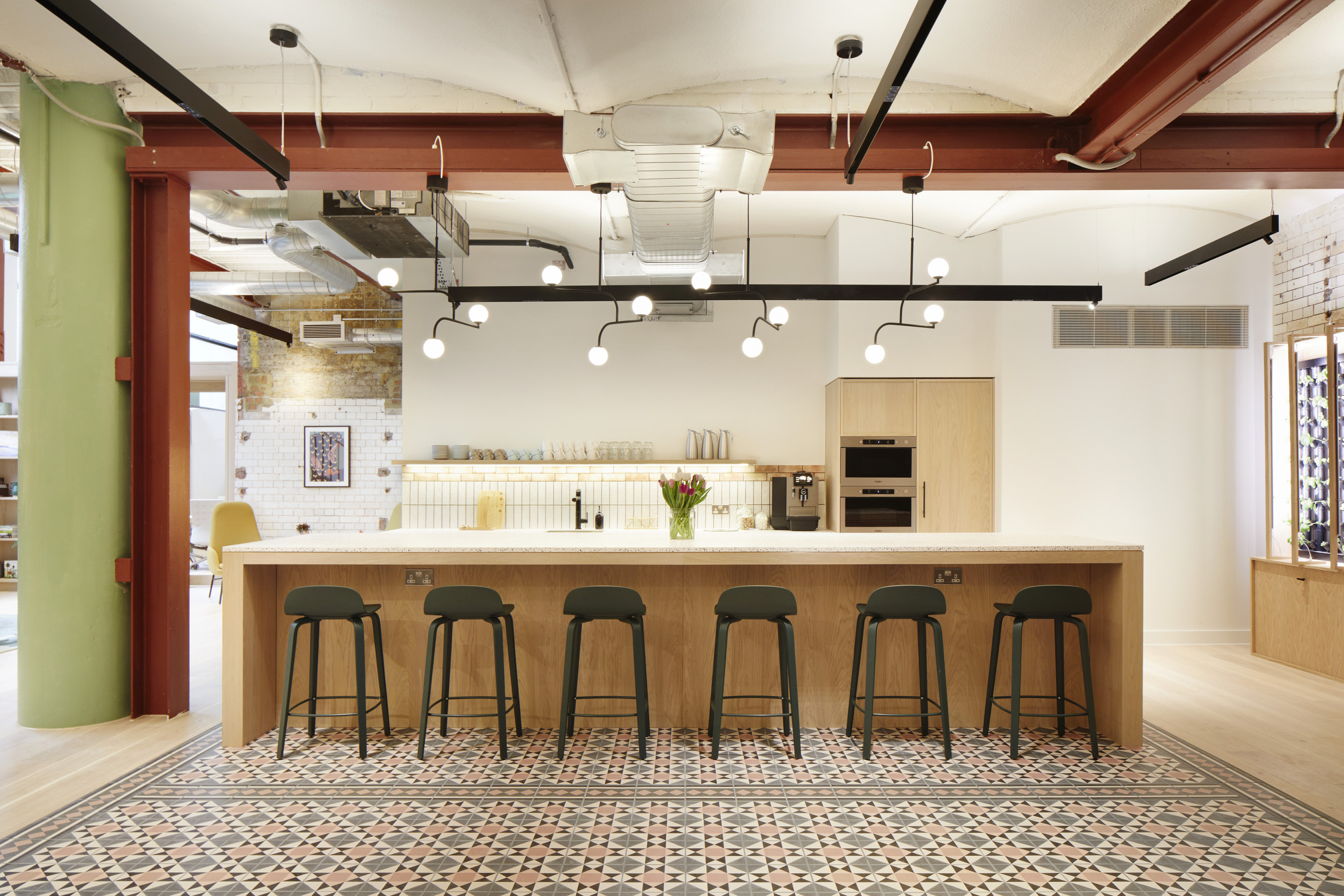 Stylish co-working kitchen at Fora Greencoat Place, designed by Modus, showcasing a central island with bar seating, geometric floor tiles, and modern lighting, providing a functional yet aesthetically pleasing space for community interaction.