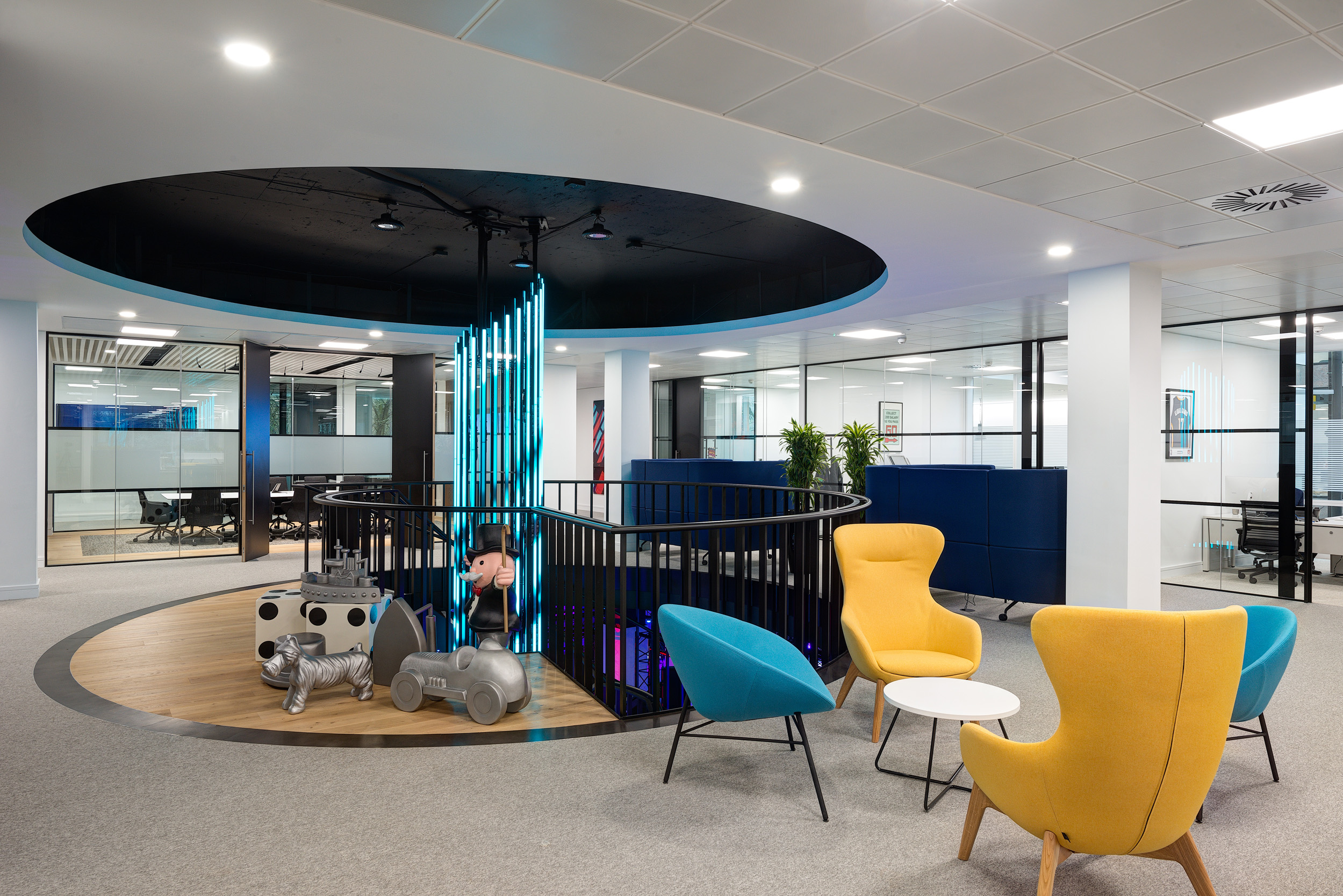 A playful and vibrant collaborative area with Monopoly-inspired figures, circular seating in bright yellow and blue chairs, and a central feature with vertical light bars.