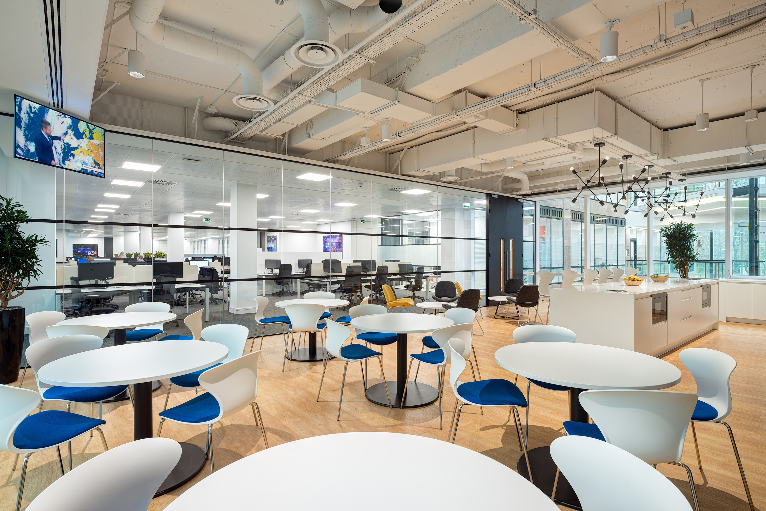 A spacious and airy cafeteria with glazing separates it from the work areas at Harman. The space has white round tables with blue accents on the chairs and modern lighting fixtures.