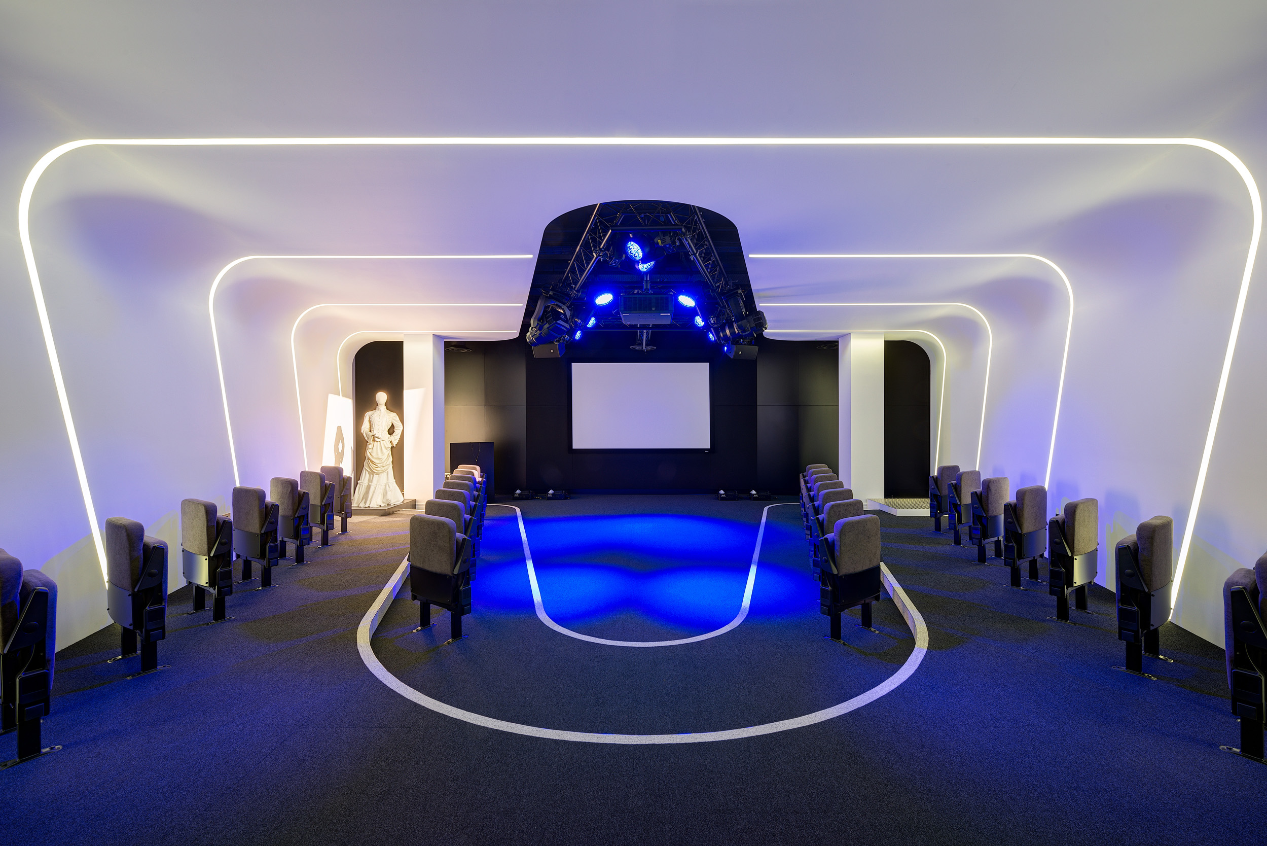 A high-tech presentation room at Harman featuring futuristic curved lighting, theatre-style seating, and an illuminated central stage area with mannequin displays.