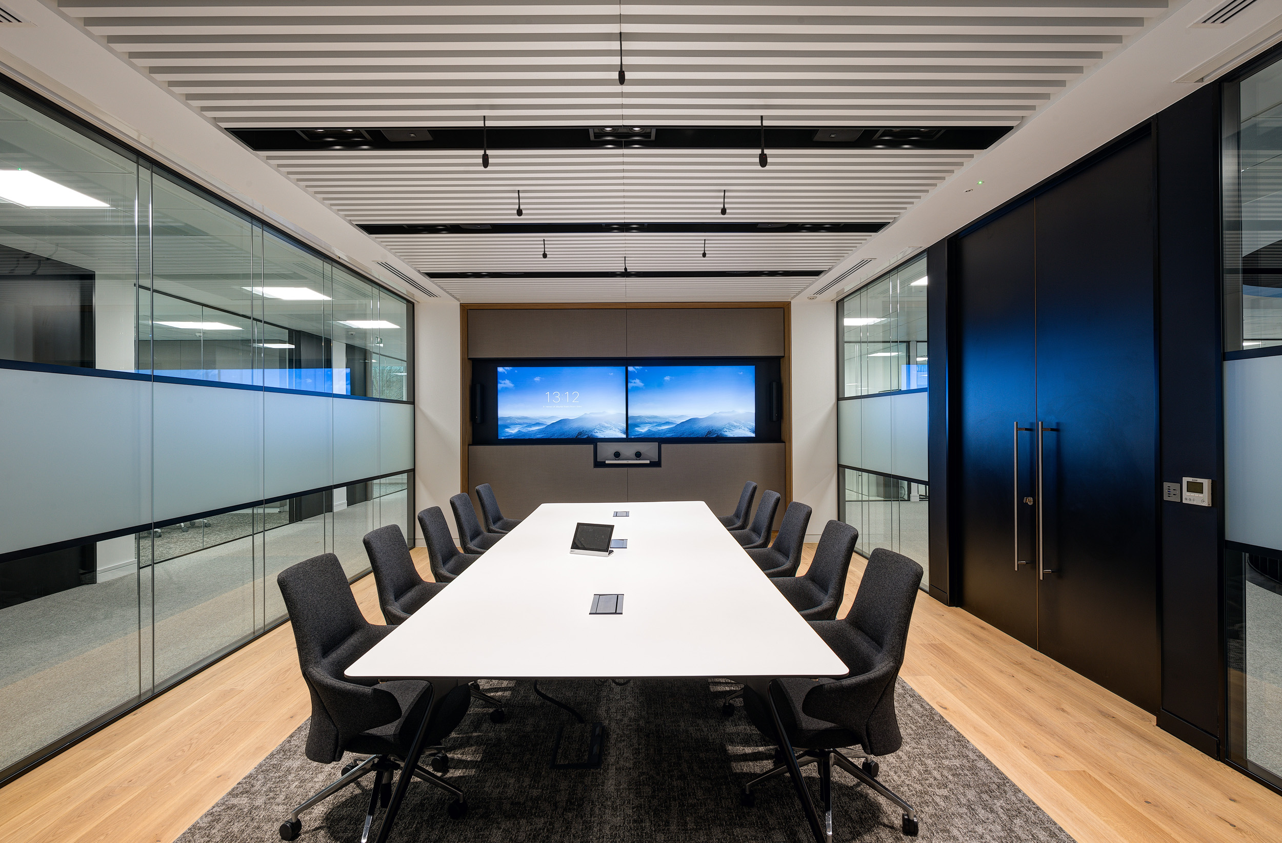 A modern conference room at Harman has a long white meeting table, grey chairs, and two large digital screens on the wall. The space is divided by glazing.