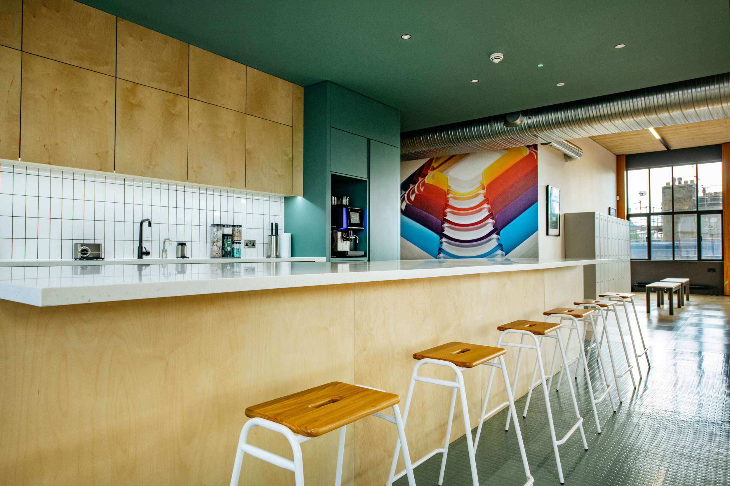 Joseph Joseph teapoint with green walls, and a large colourful painting. High white stools, and white tiles on the teapoint.