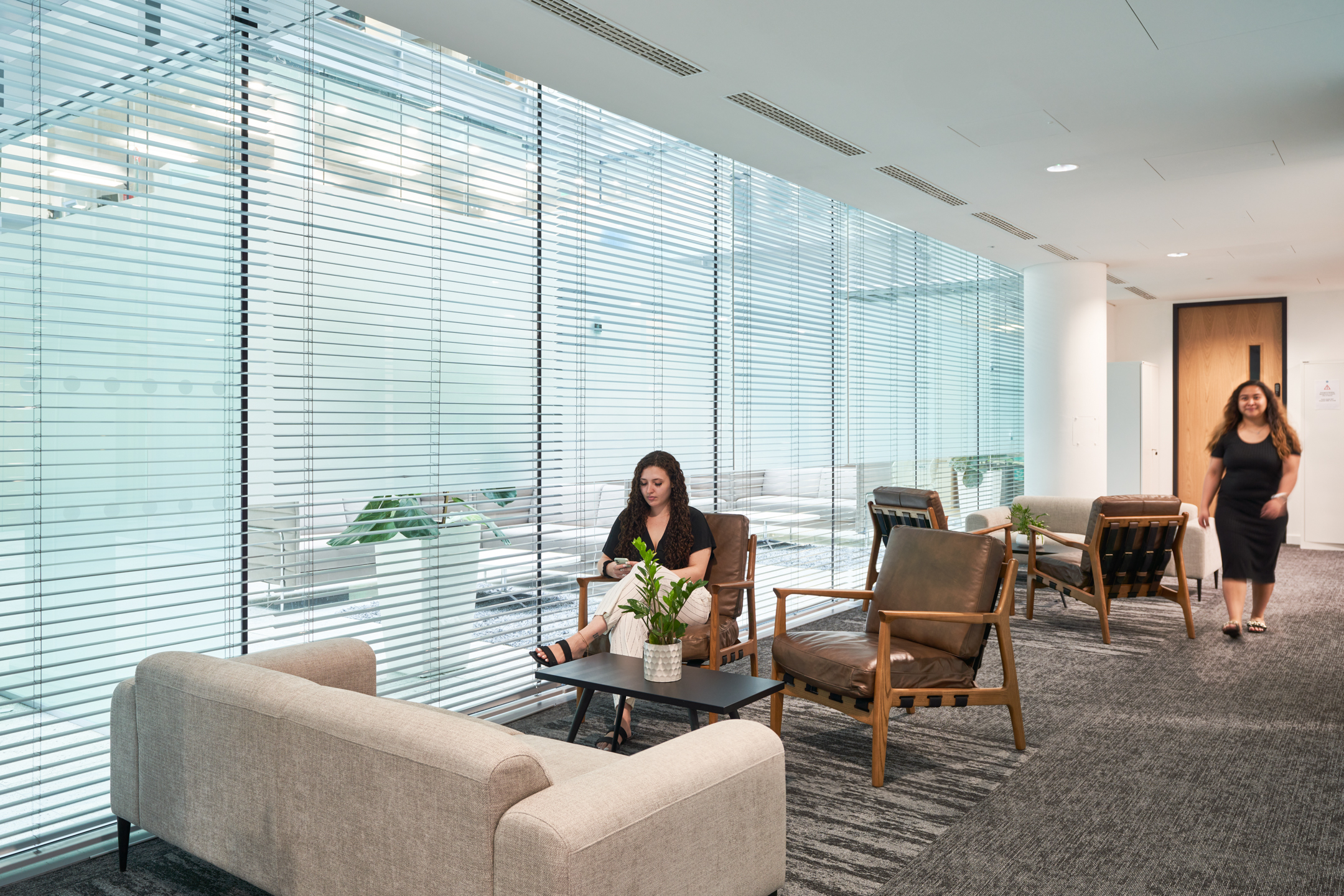 Two women are seated in a well-lit, glass-walled seating area, featuring contemporary armchairs and a soft grey sofa, providing a tranquil space within LGC's 100 Victoria Street office, designed by Modus Workspace.