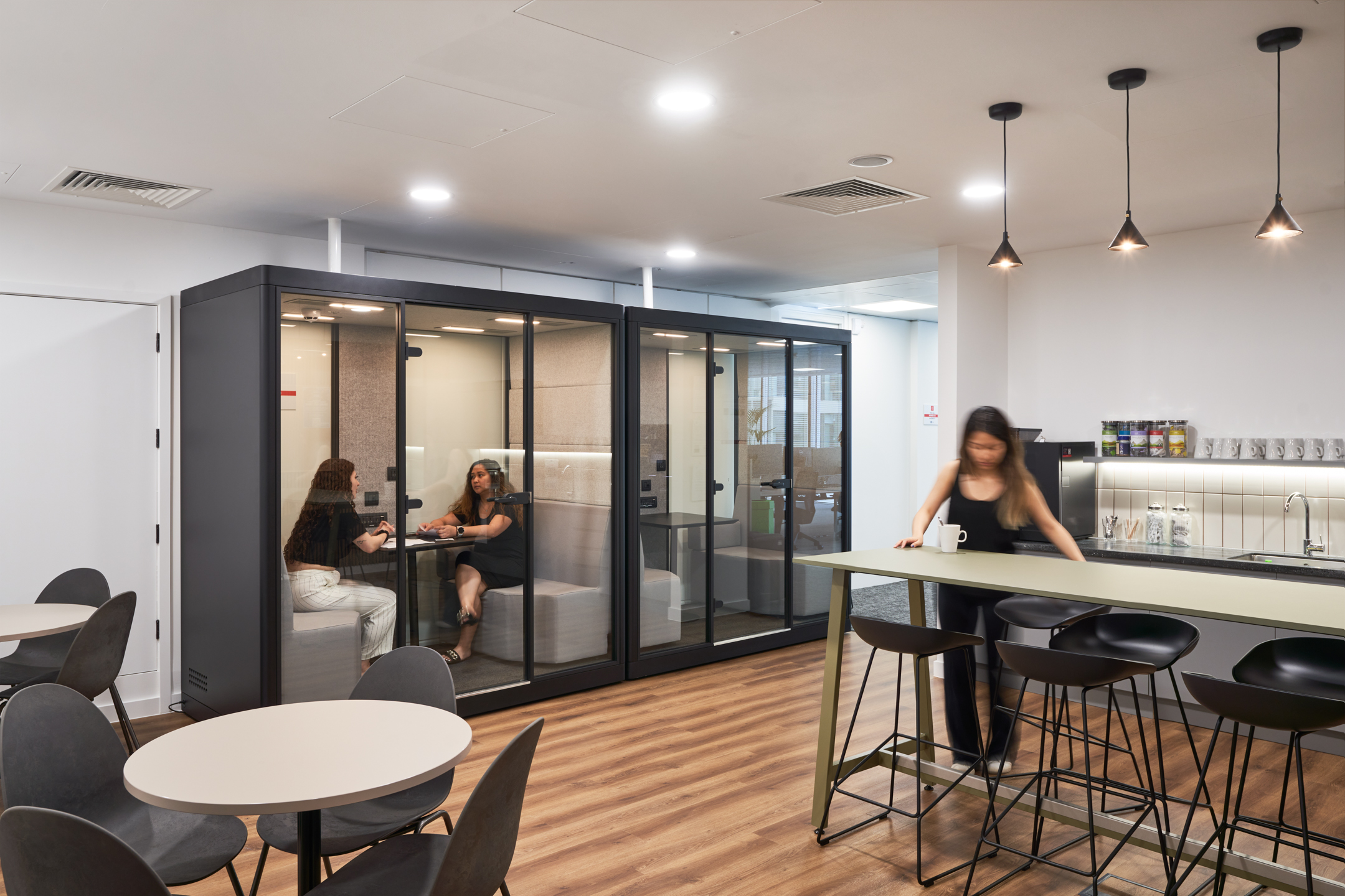 A teapoint area with sleek black chairs and a high counter is adjacent to acoustic meeting pods, where two women are engaged in a discussion. This setup within LGC's 100 Victoria Street office, designed by Modus Workspace, promotes collaboration and informal meetings.