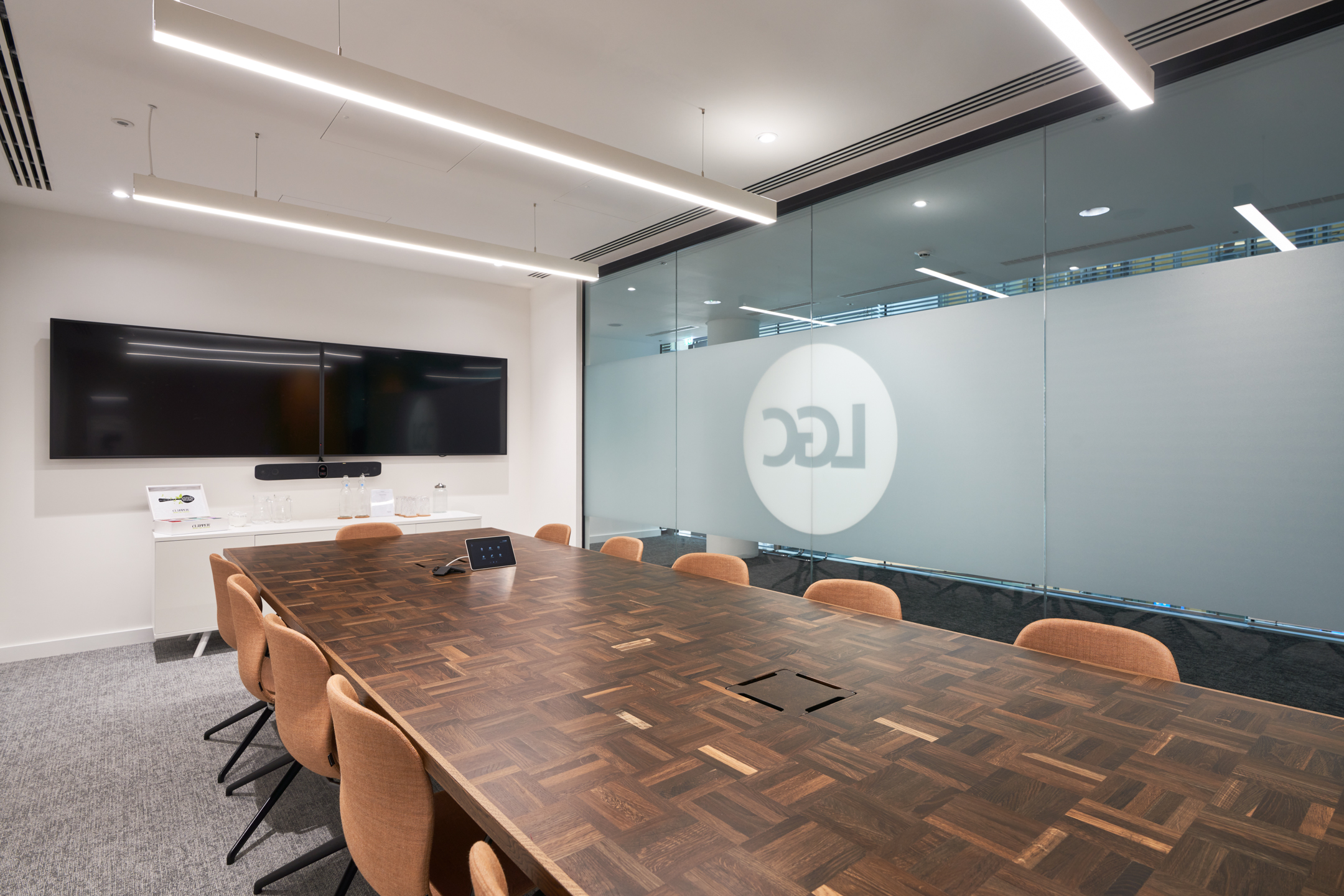 A modern meeting room at LGC's 100 Victoria Street office, designed by Modus Workspace, features a wooden table with a parquet design, comfortable seating, and dual screens for presentations and video conferencing.