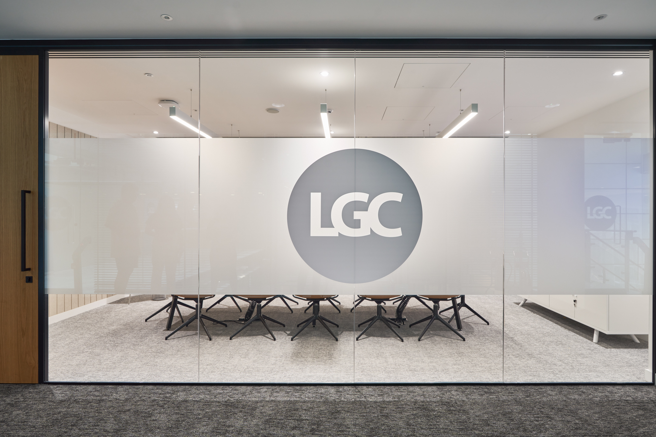A glass-walled meeting room at LGC's 100 Victoria Street office, prominently displaying the LGC logo on frosted glass. The minimalist design by Modus Workspace ensures privacy while maintaining an open feel.