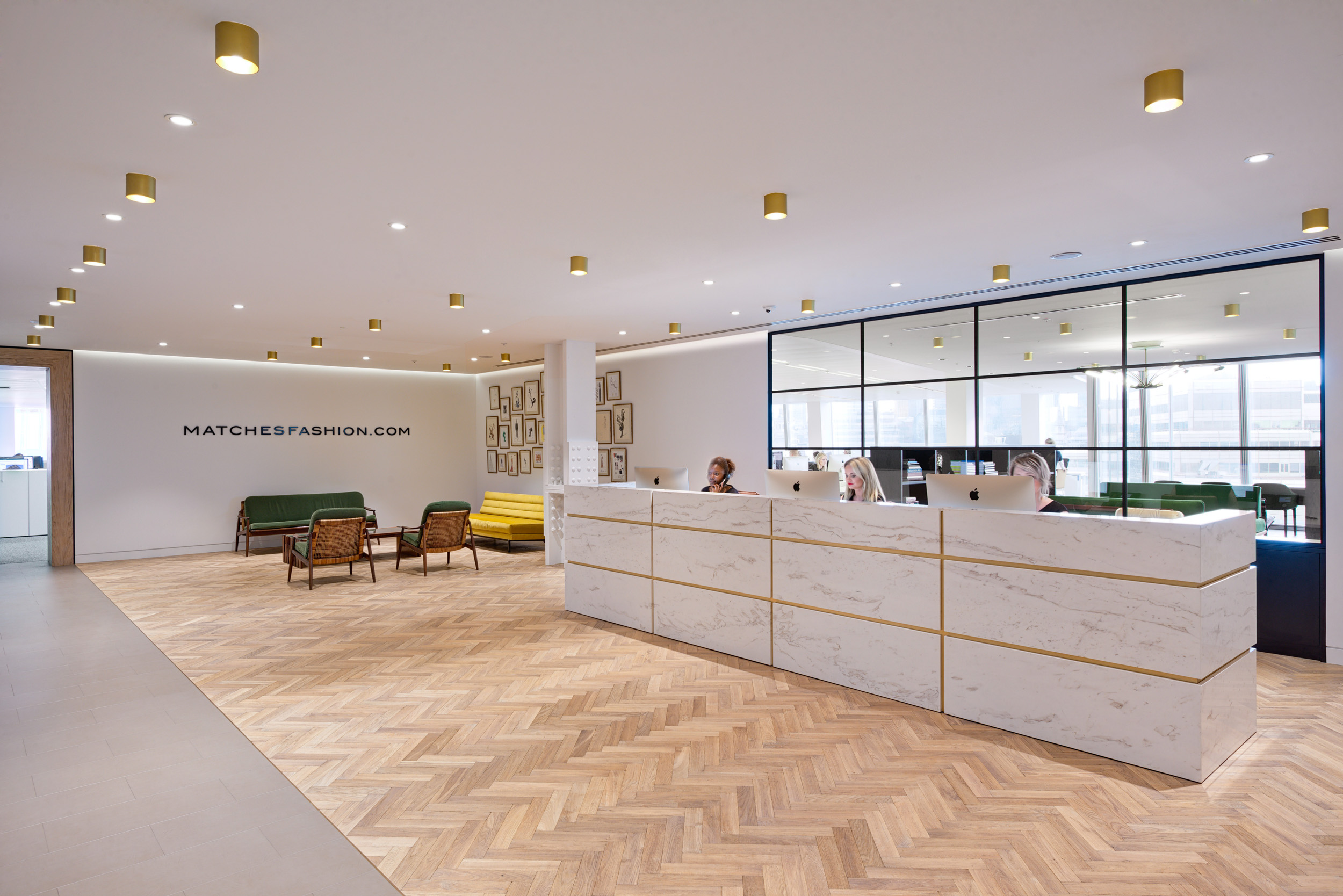 A modern open-plan office at Matches Fashion, designed by Modus Workspace, where employees are seen working at their desks. The clean, minimalist design is complemented by a stylish staircase leading to an upper level, creating a cohesive workspace.