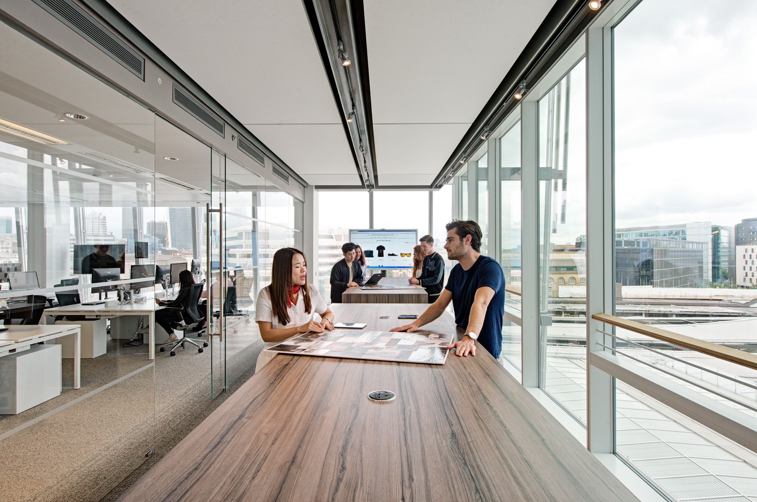 A spacious teapoint at Matches Fashion, designed by Modus Workspace, showcasing a communal wooden table and modern seating, with employees enjoying the open and airy environment. The industrial ceiling adds to the contemporary feel.