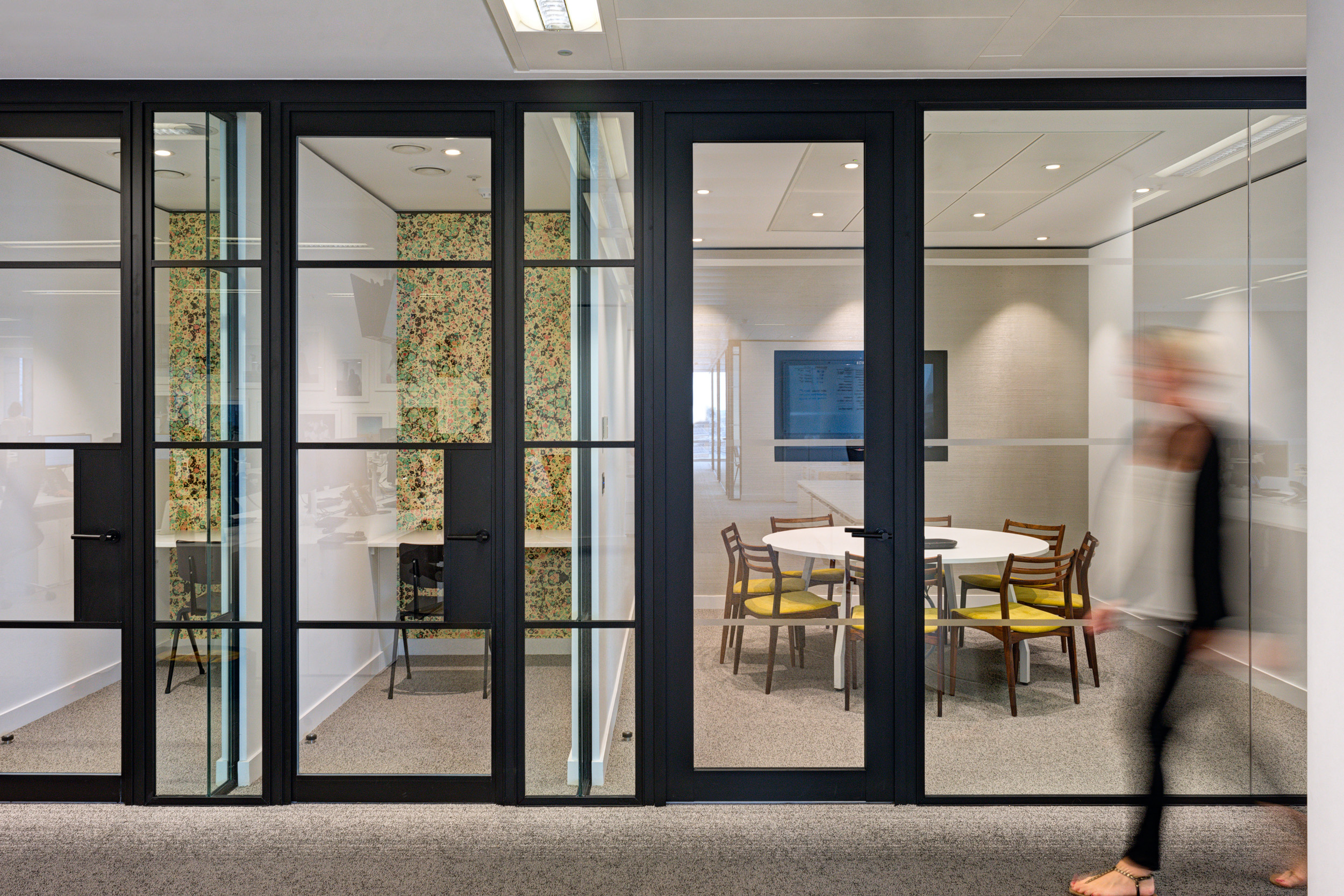 A collaborative workspace at Matches Fashion, designed by Modus Workspace, featuring large windows offering natural light and views of the city. Employees are engaged in discussions around a table, emphasising teamwork in a bright, modern setting.