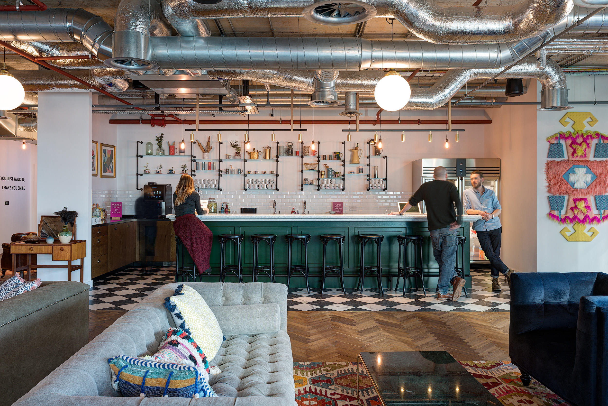 A colourful and industrial-inspired bar space with green cabinetry and exposed ceiling pipes at Mindspace 144 Whitechapel, designed by Modus Workspace.