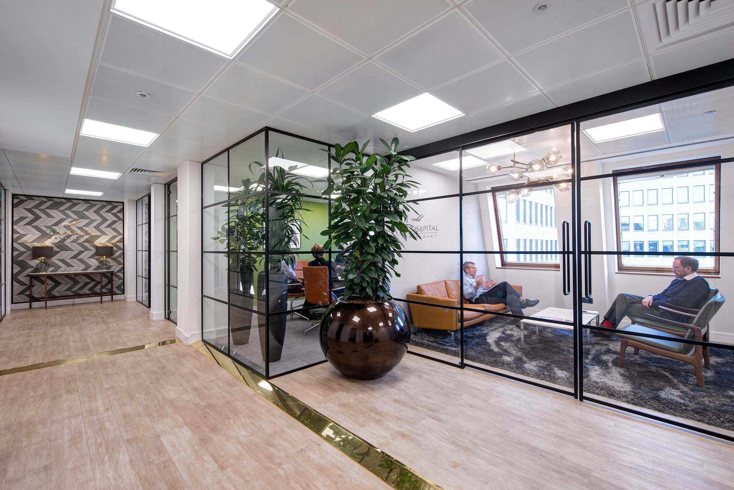 The corridor of Otus Capital Management, designed by Modus Workspace, lined with glass-walled meeting rooms, allowing for a flow of natural light, with employees seen engaging in discussions, reflecting the collaborative workspace design.