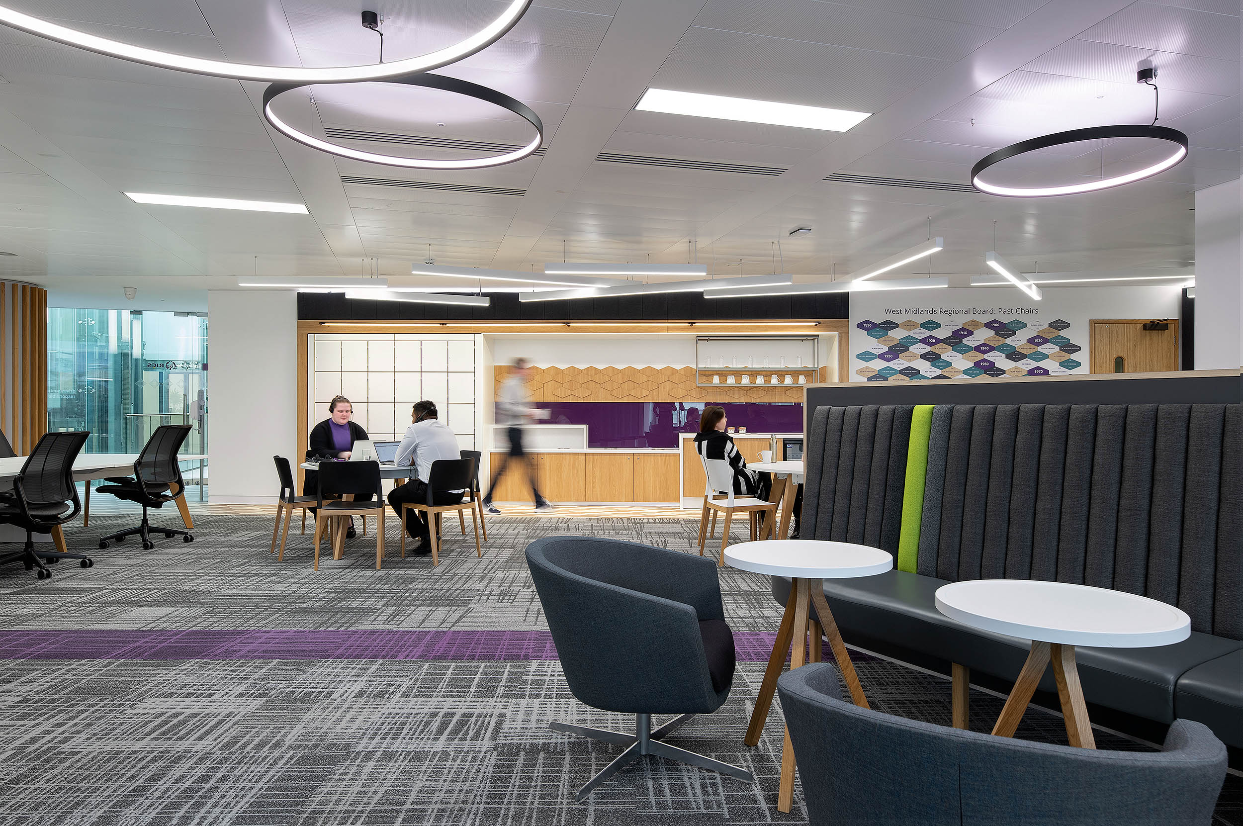 Open-plan teapoint at RICS featuring black chairs, wooden tables, overhead circular lighting, and a seating area with a purple accent wall.