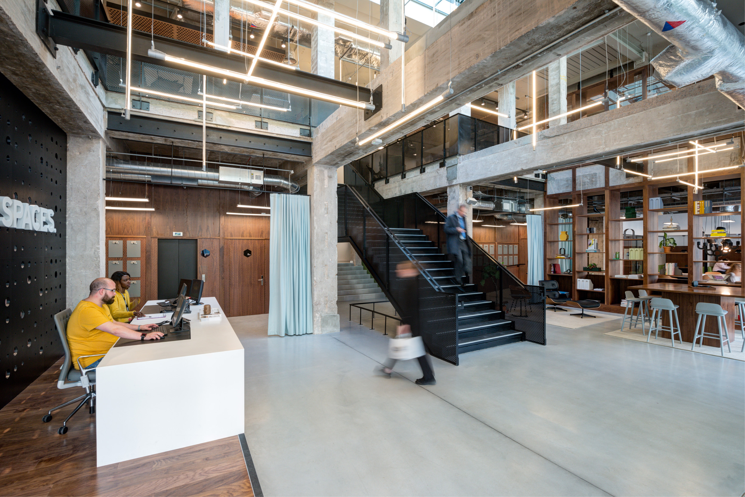 View of the reception area and open workspace at Spaces Peter House, showcasing a black metal staircase and wooden shelving.