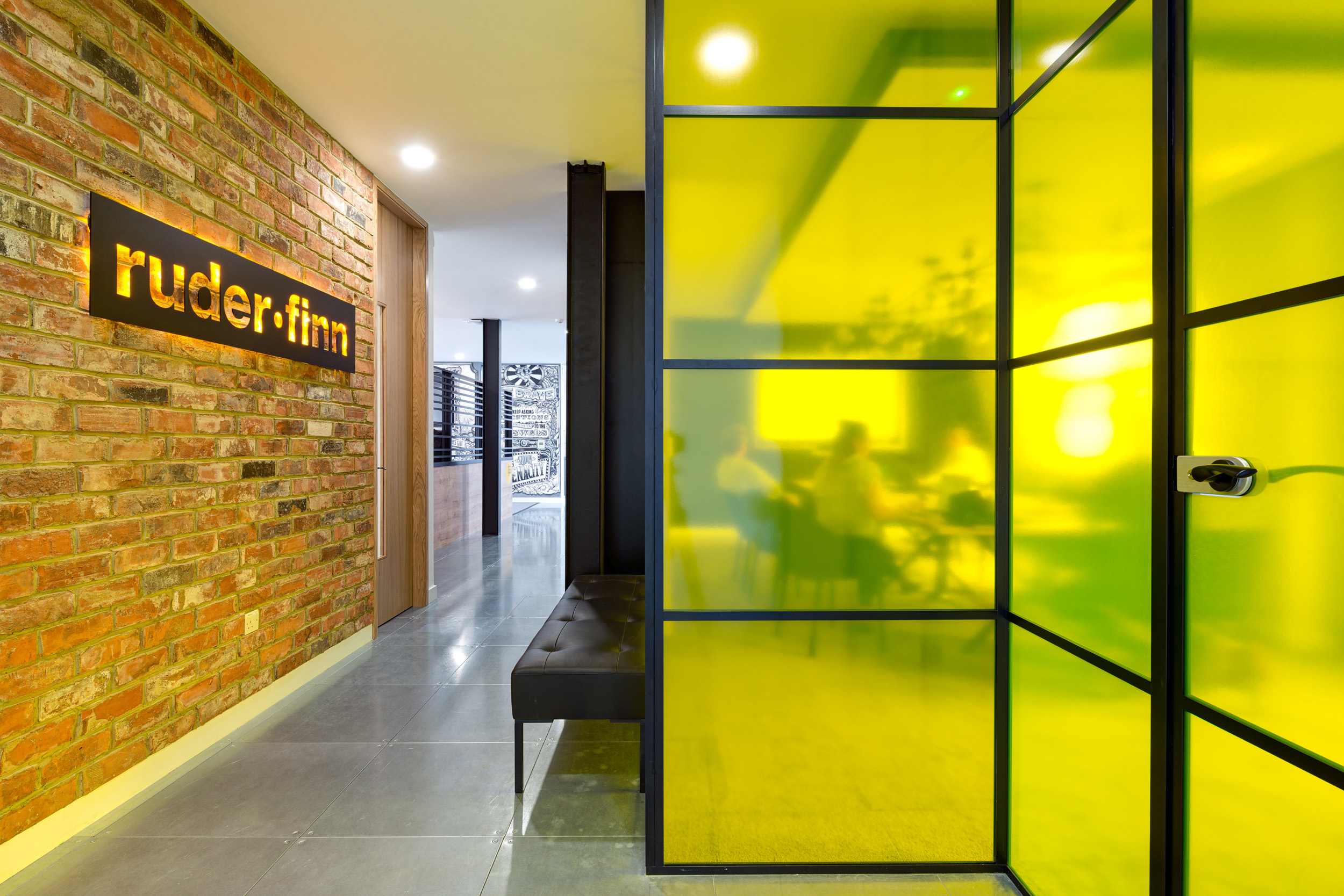 The Ruderfinn office entrance, designed by Modus Workspace, features an illuminated company sign on a brick wall, with a modern, yellow-accented glass partition.