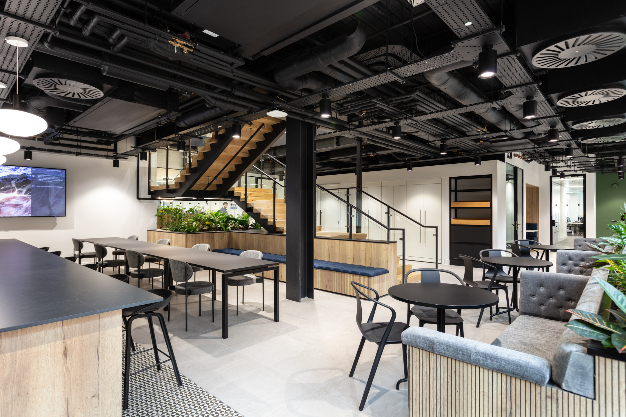 A sleek, open-plan tea point and collaboration area at Russell Reynolds featuring black industrial ceilings, wooden accents, and ample seating areas. Designed by Modus Workspace, the space includes greenery and a staircase leading to an upper level, providing a dynamic environment for relaxation and collaboration.
