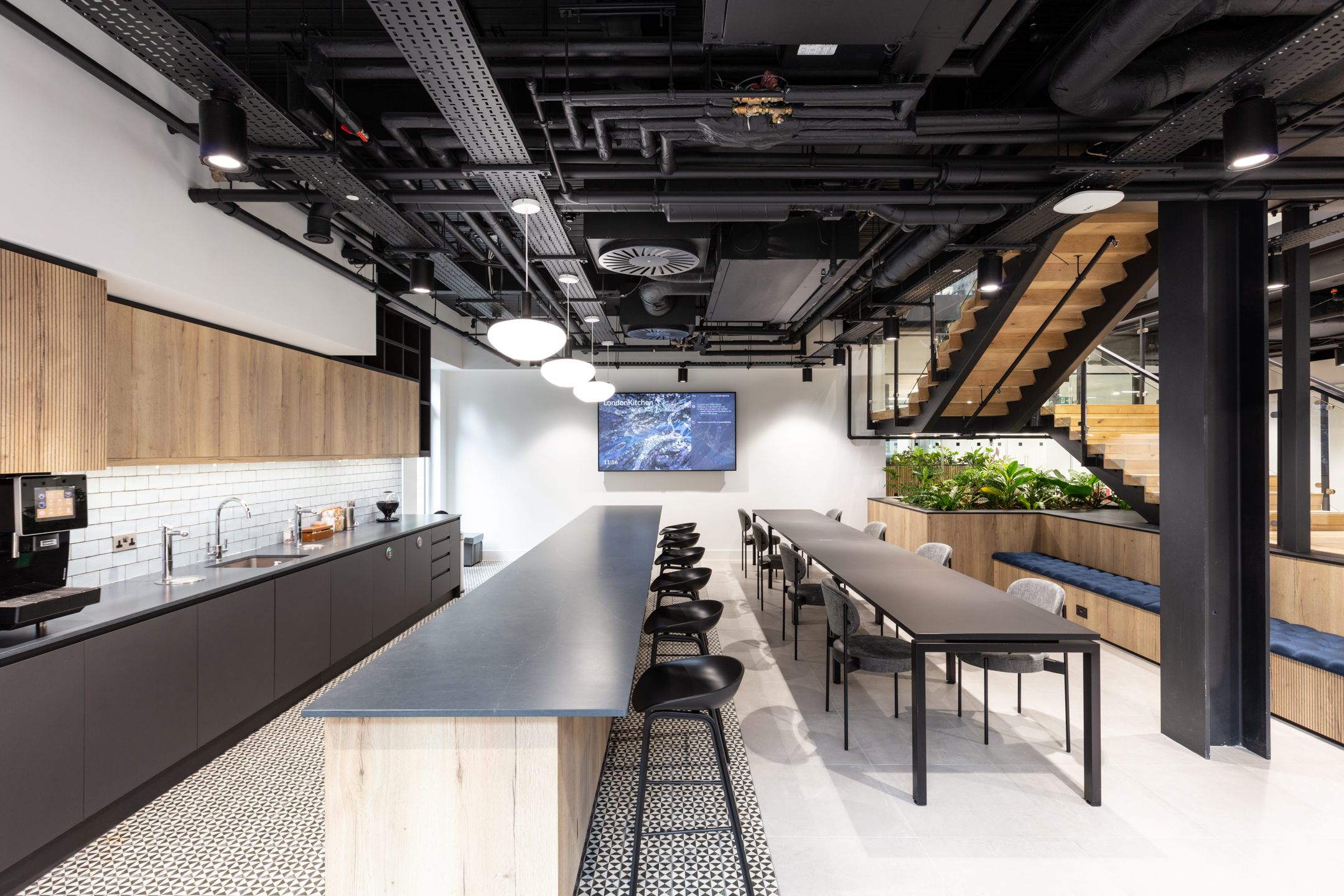 The teapoint area at Russell Reynolds, designed by Modus Workspace, showcases a minimalist aesthetic with light wood cabinetry, black countertops, and white tiled backsplashes. The seating space is designed for comfort with long communal tables, perfect for team gatherings.