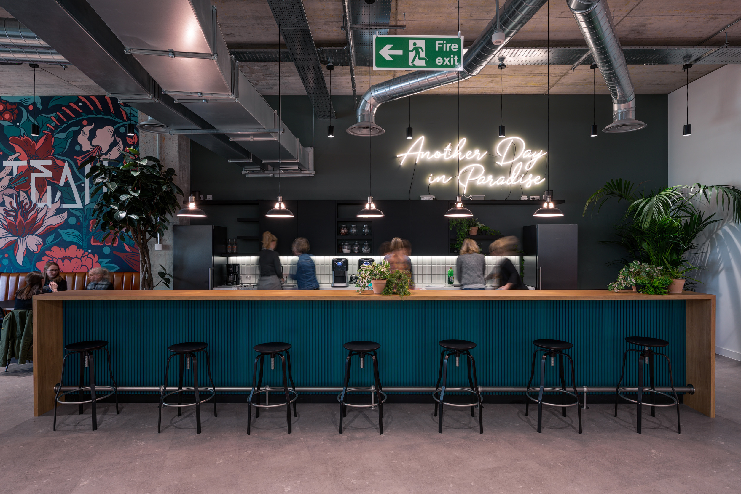 Spacious modern office kitchen and bar area at SLG, designed by Modus. The space features a long teal bar counter lined with black stools and a vibrant neon sign saying 'Another Day in Paradise' against a dark grey wall. Lush greenery and industrial-style lighting enhance the lively ambiance