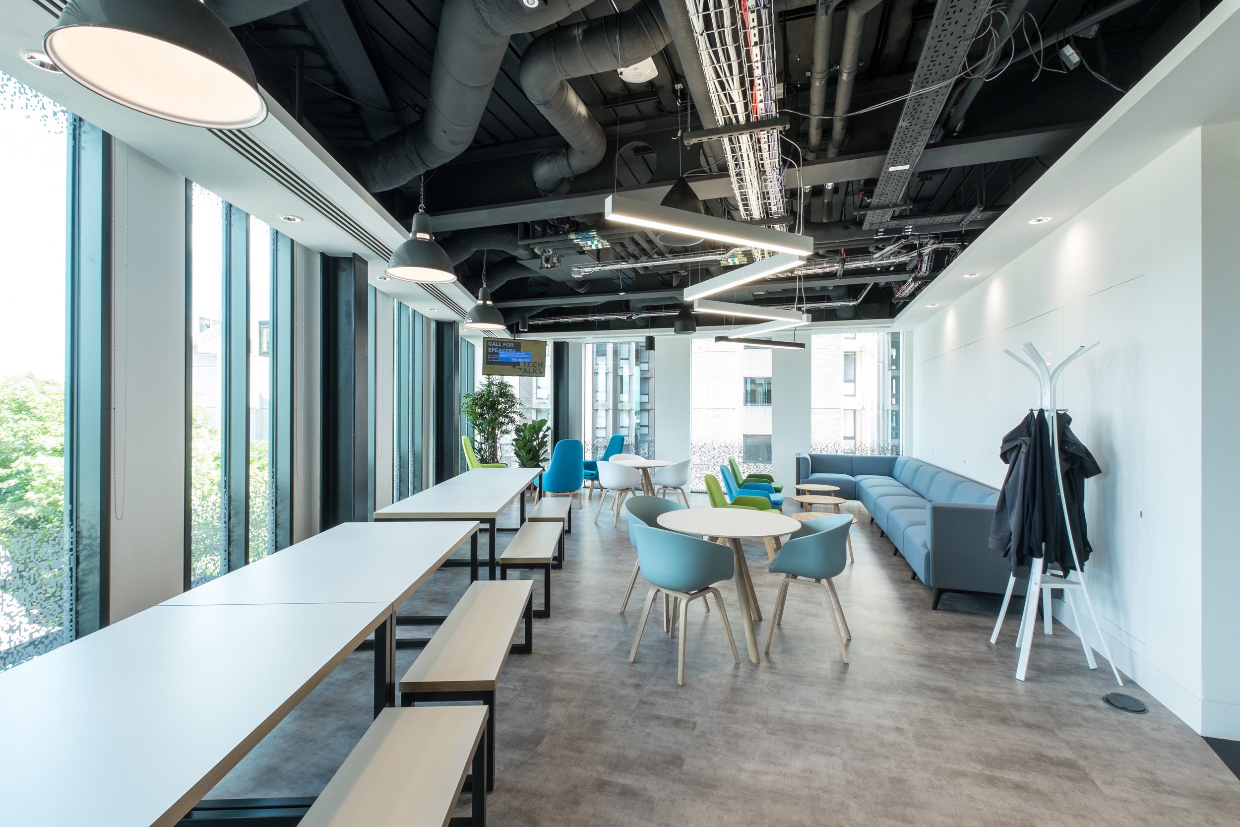A versatile seating area in the Skyscanner office with a mix of benches and casual chairs, large windows, and an industrial ceiling with exposed ductwork.