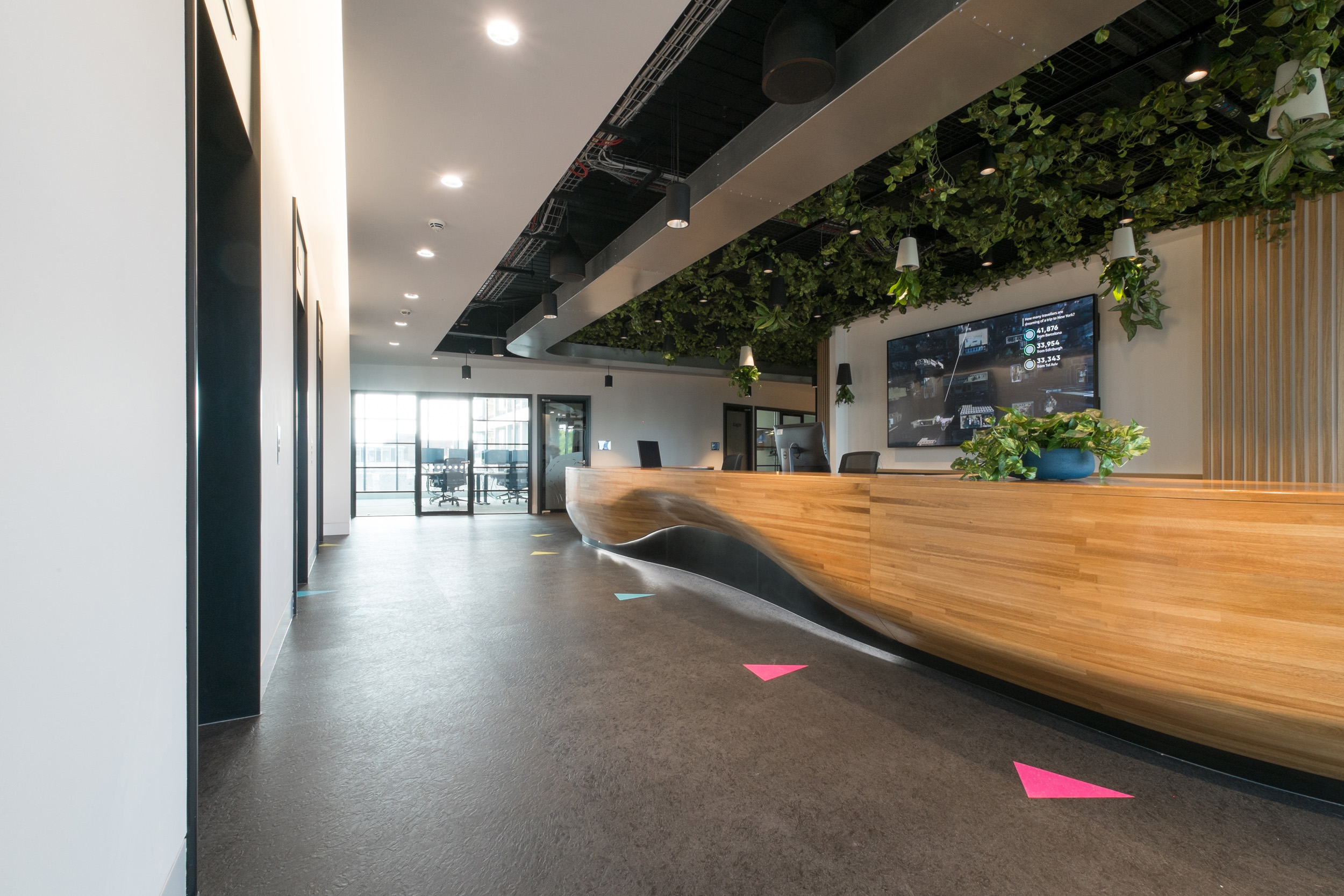 The reception desk at Skyscanner’s office has a wood-accented counter, overhead plants, and a digital display framed by overhead greenery and modern lighting.