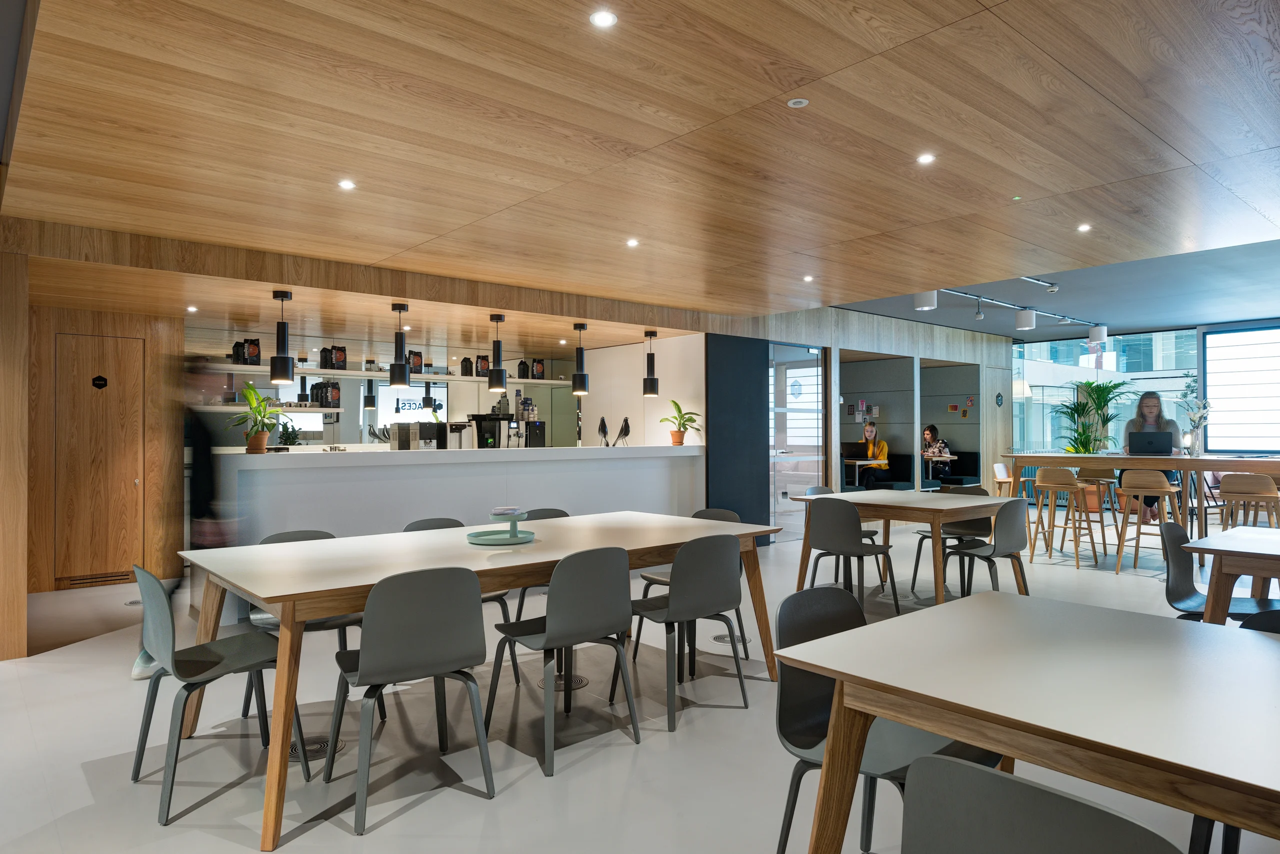 Breakout area and workspace at Spaces Uxbridge with large wooden tables, pendant lighting, and a sleek teapoint area in the background. Employees are seen using various seating arrangements.