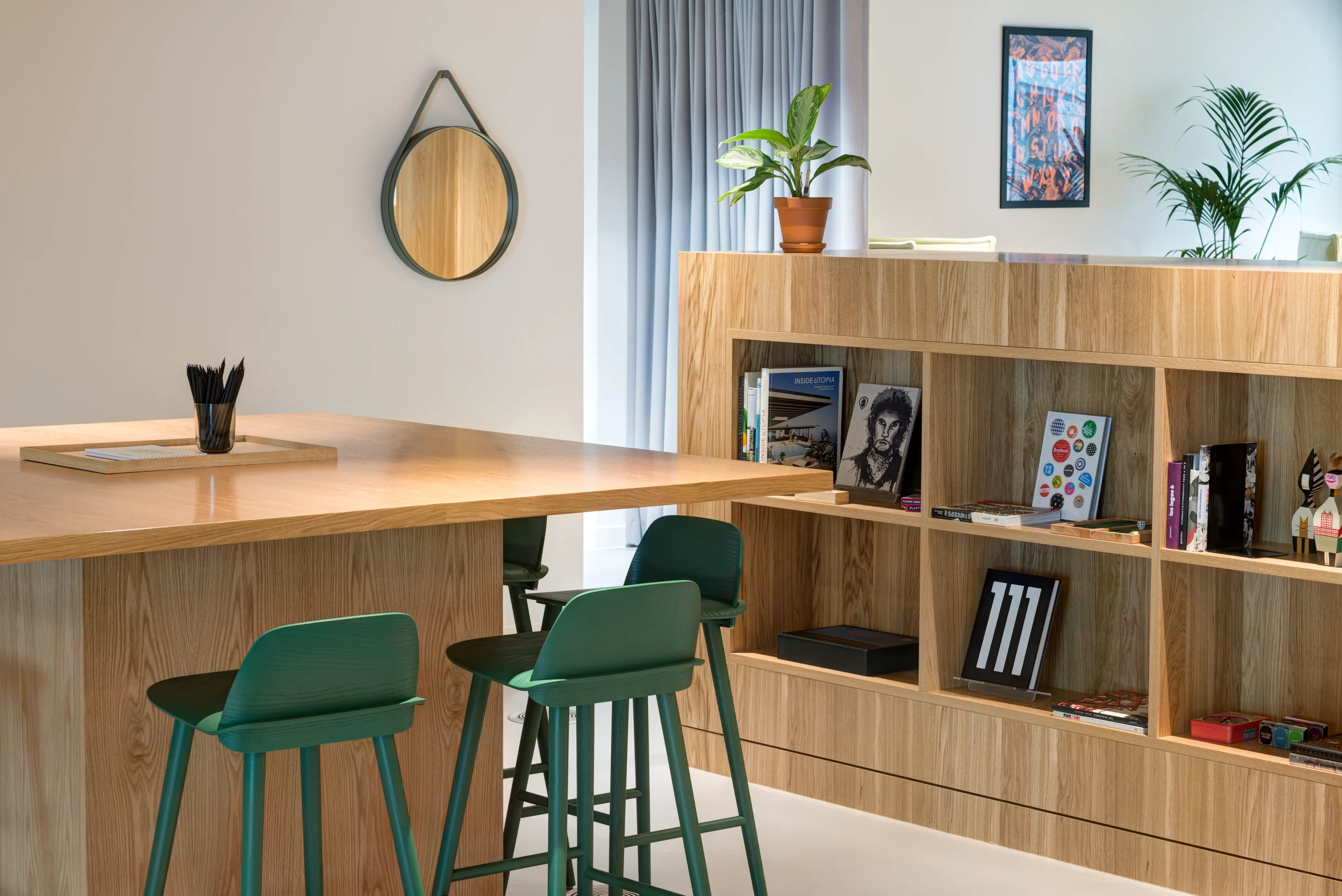 A high wooden counter with green bar stools at Spaces Uxbridge, complemented by shelves filled with books and decorative items. A mirror and potted plants add to the ambience.