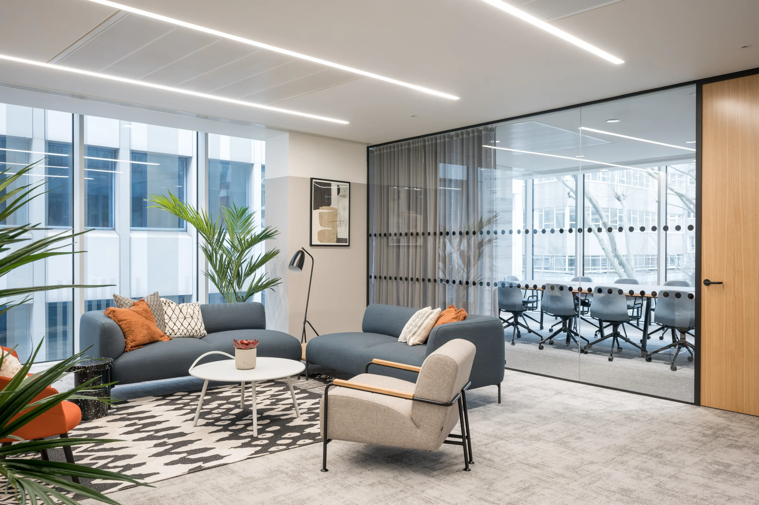 A relaxed office lounge area with blue sofas and various chairs around a central coffee table, positioned on a geometric patterned rug. The room includes a glass-walled meeting area in the background, illustrating Modus's modern design for State Street Global Advisors.