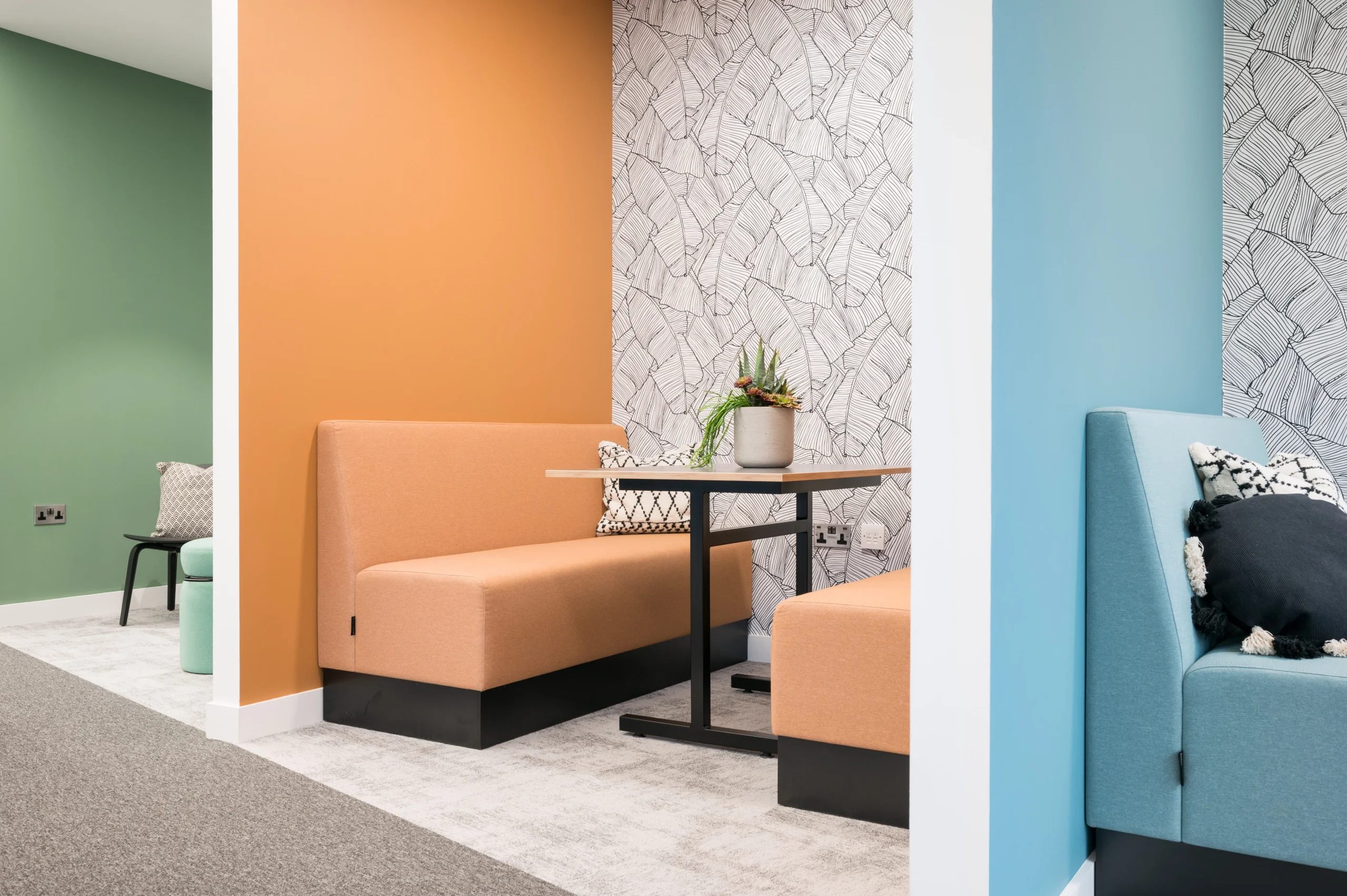 A colourful nook in the office with orange and teal seating against a wall with a geometric leaf pattern, highlighted by a smooth wooden table in the middle. The area is part of Modus's interior design for State Street Global Advisors.
