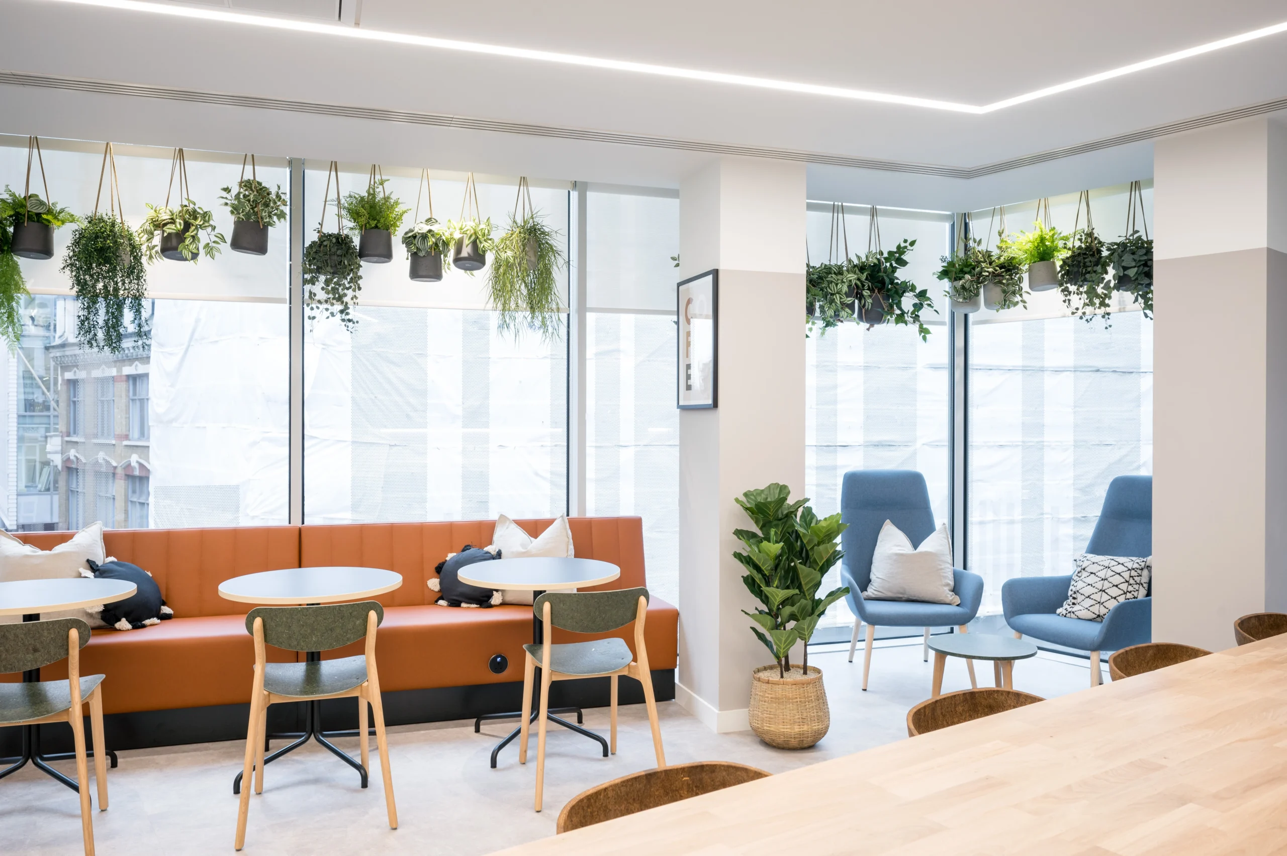 Another angle of the office breakout area showing orange and blue seating and round tables. The space is bright with natural light filtering through large windows, complemented by green hanging plants above and white, geometric lighting. Designed by Modus for State Street Global Advisors.