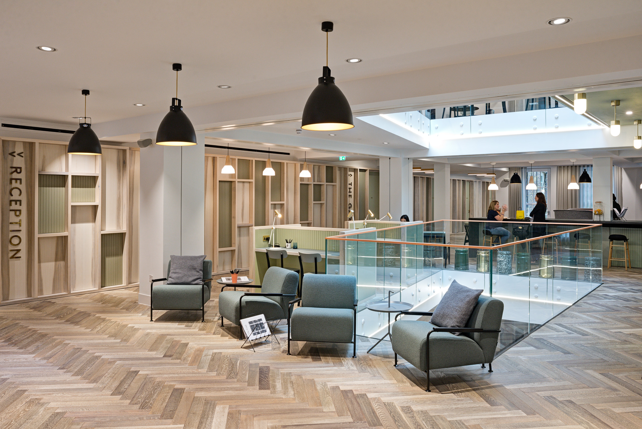 The Office Group Wimpole Street reception and waiting area, designed by Modus Workspace, with stylish pendant lighting and modern furnishings. People are seen working in various seating areas, including a central reception and upper-floor desks.