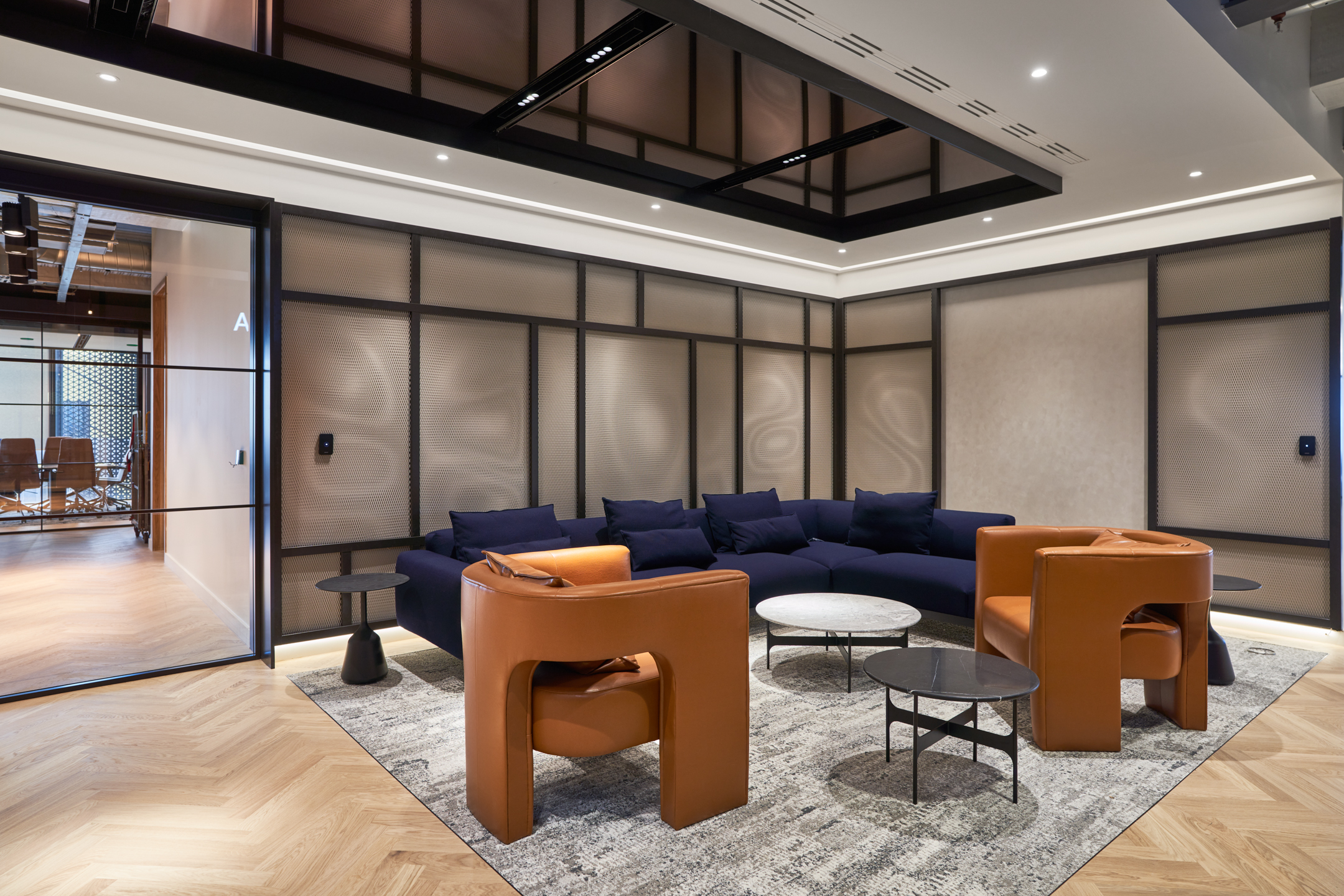A casual lounge area at United Talent Agency designed by Modus offers a relaxed seating arrangement with dark blue sofas and stylish orange chairs. The room includes wooden flooring, geometric patterns in the furniture layout, and a serene, welcoming ambiance for informal meetings.