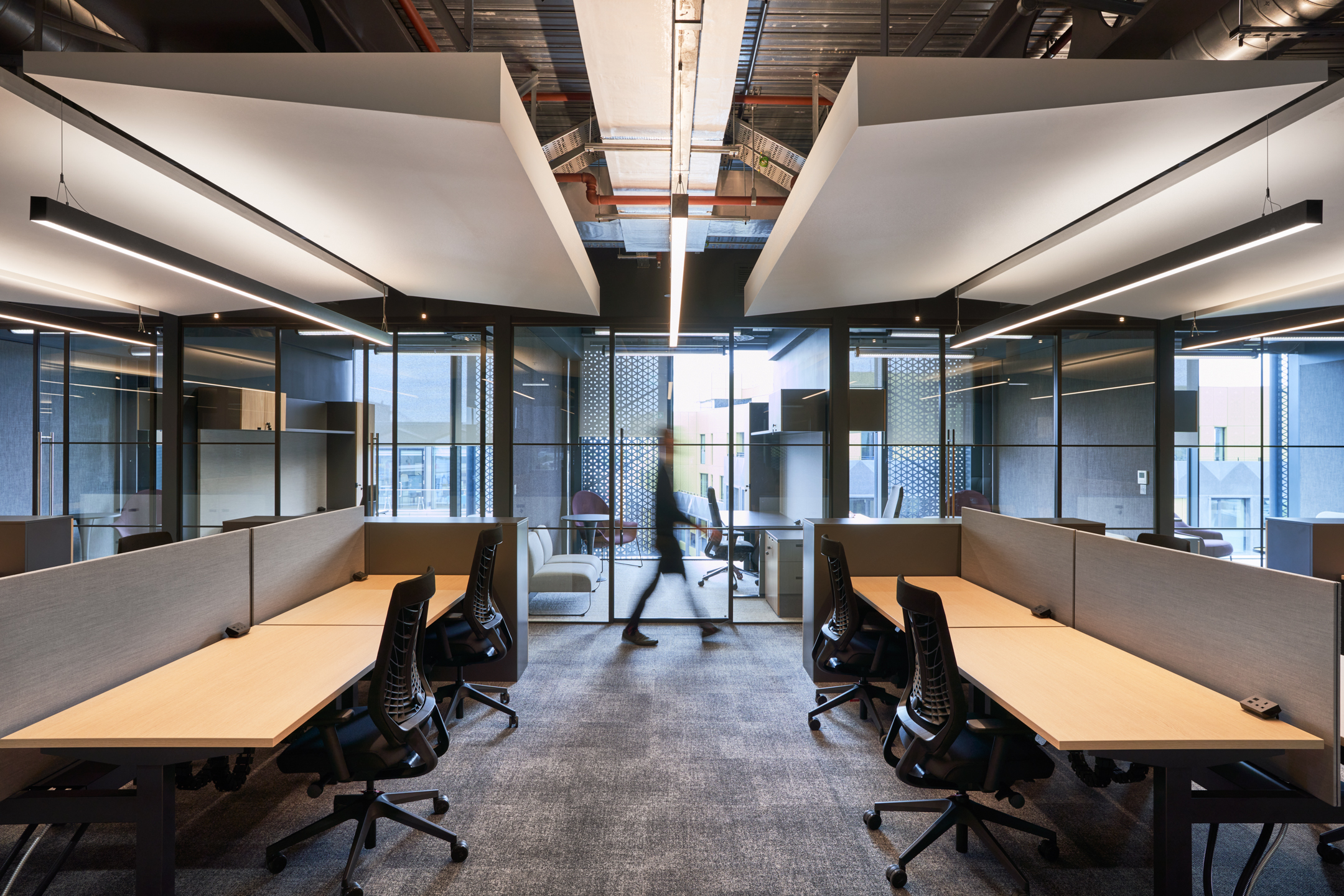 This open workspace at United Talent Agency, designed by Modus, features angular light fixtures and contemporary furniture. The space is arranged to foster collaboration, with a mix of enclosed glass meeting rooms and open desk areas that promote a dynamic work atmosphere.