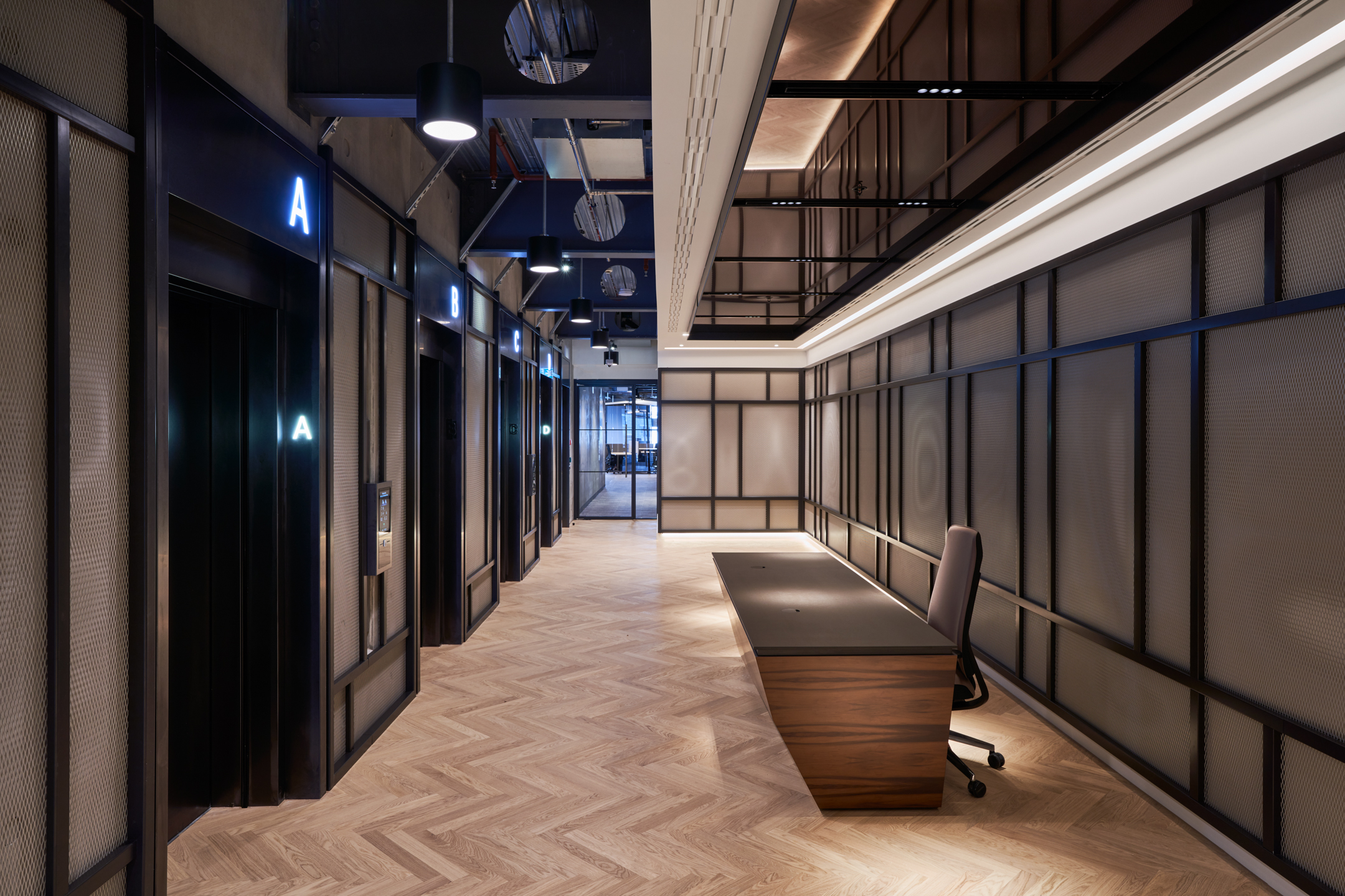 A modern and elegant hallway at United Talent Agency showcases sleek office pods with glass doors marked by illuminated letters. The design by Modus features dark tones, herringbone wood flooring, and spot lighting, providing a stylish yet functional workspace environment.